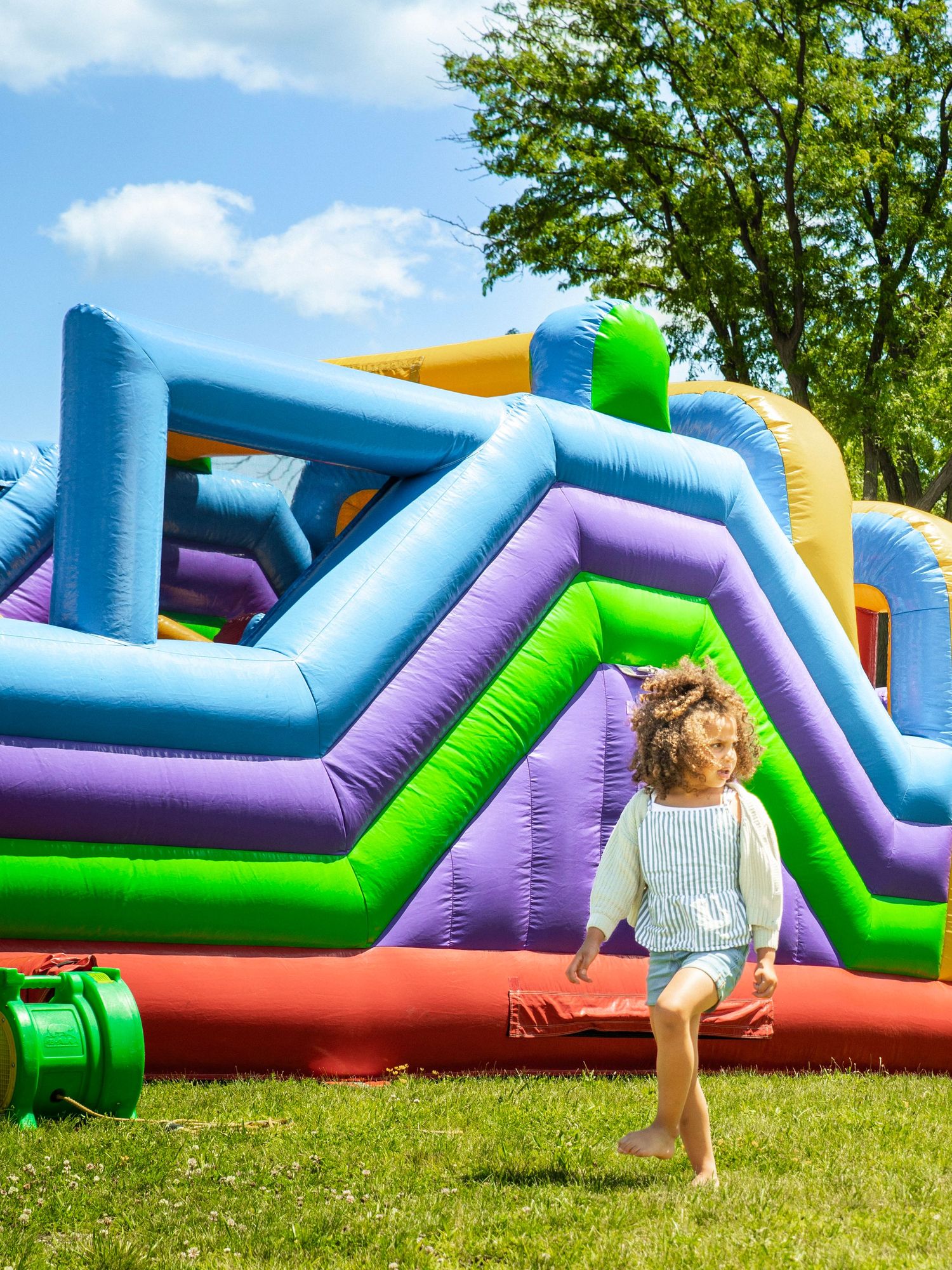 A young child with curly hair runs joyfully on green grass beside a colorful inflatable bounce house obstacle course.