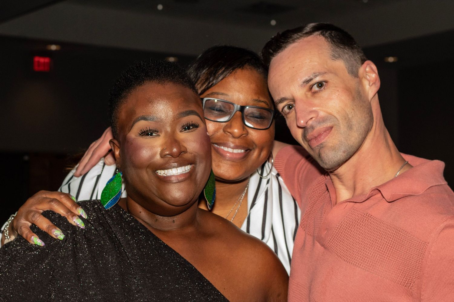 Three friends smiling and posing together at an indoor event, two women and one man in casual attire.