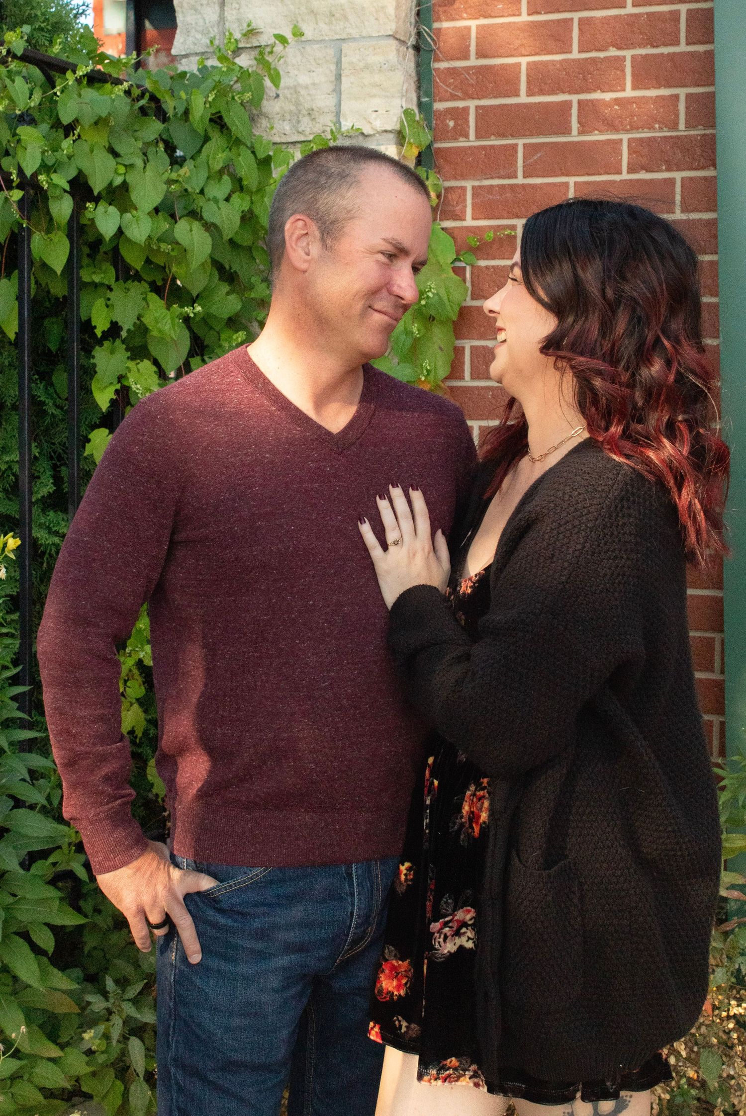 A couple in burgundy and black share a tender moment against a brick wall with green ivy.