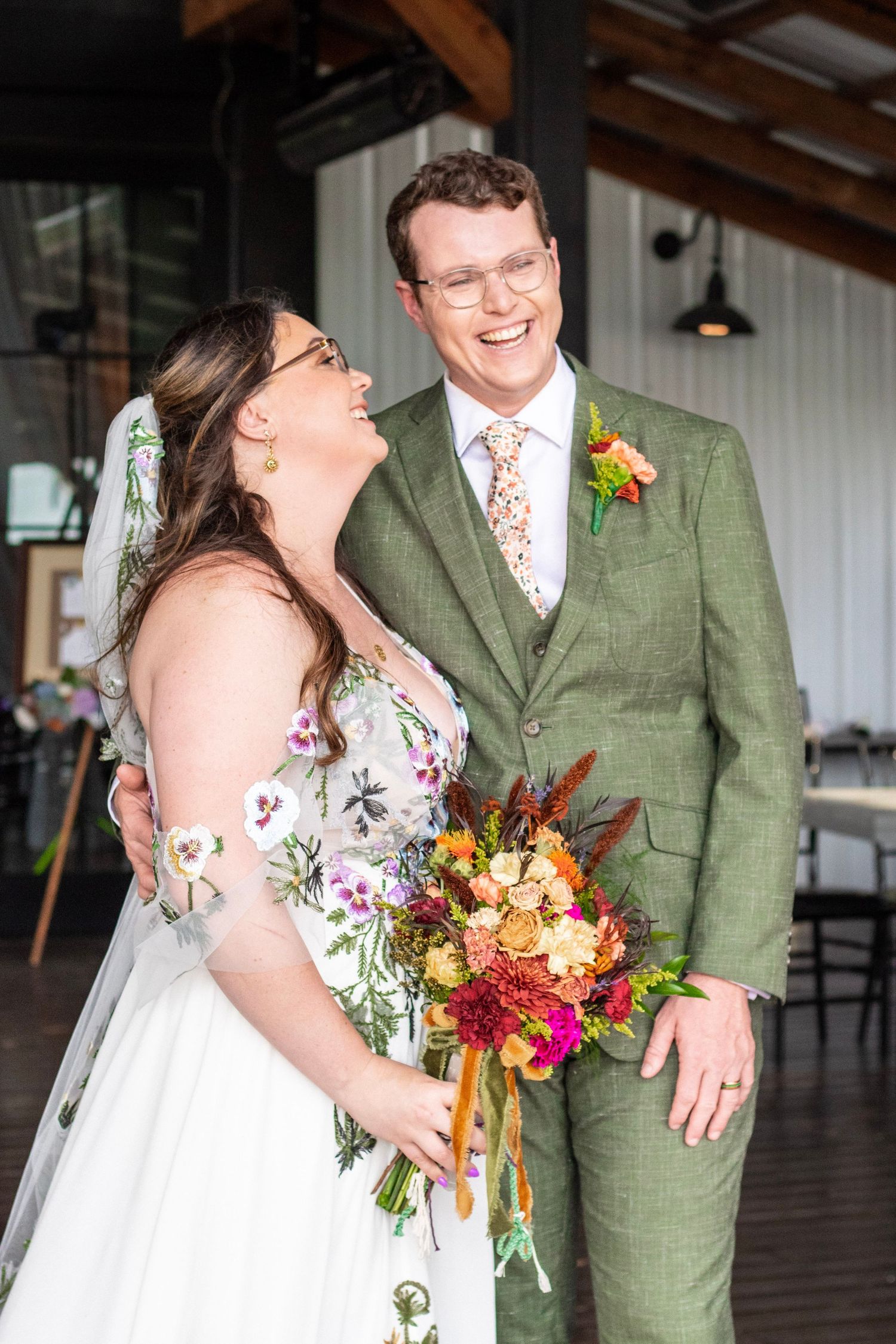 Laughing bride in floral gown and groom in green suit holding colorful bouquet at indoor wedding venue.