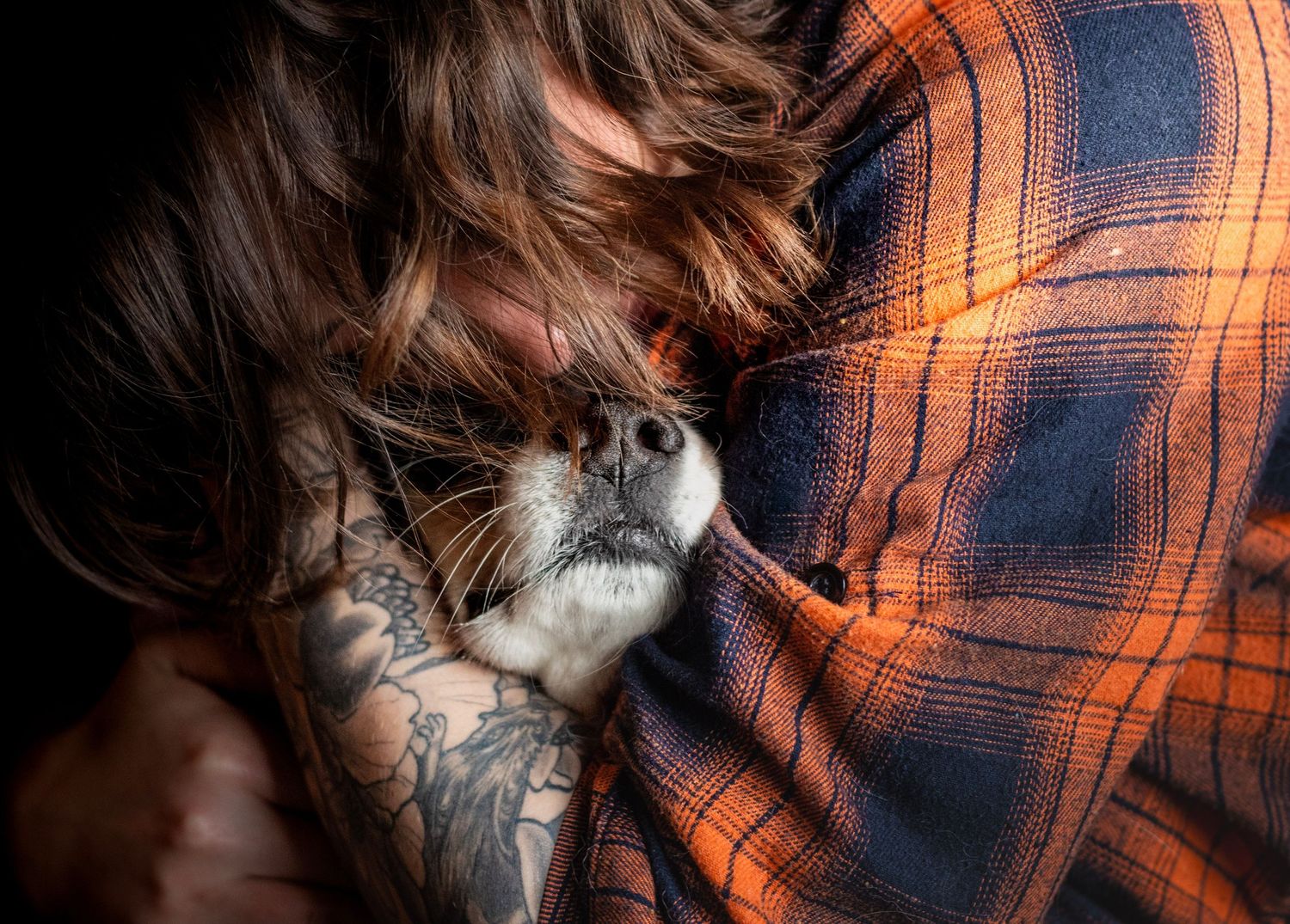 Tattooed person in orange plaid shirt cuddling a small white cat, with hair falling over their face.