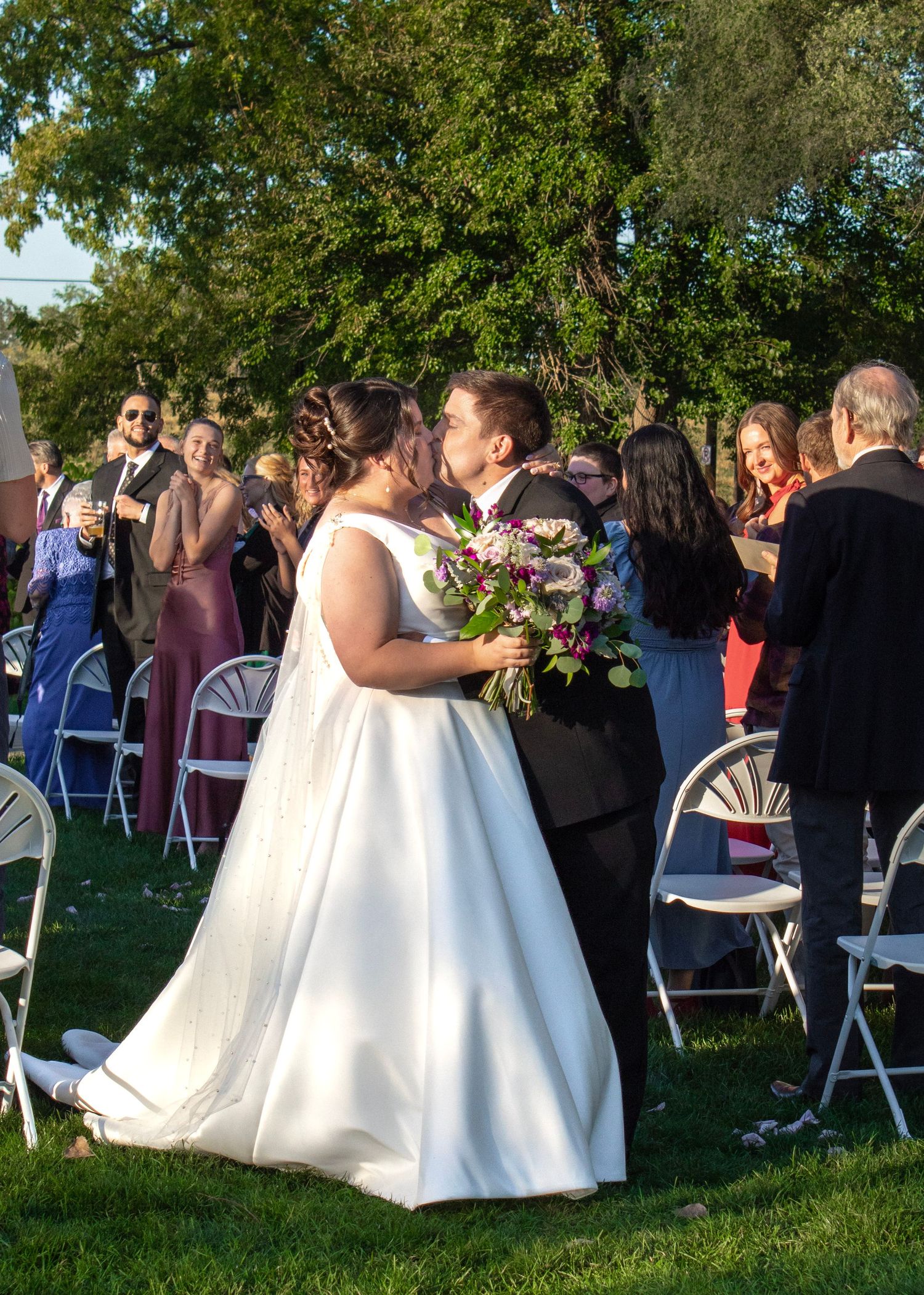 Bride and groom share a kiss at outdoor wedding ceremony, surrounded by guests on green lawn with trees in background.