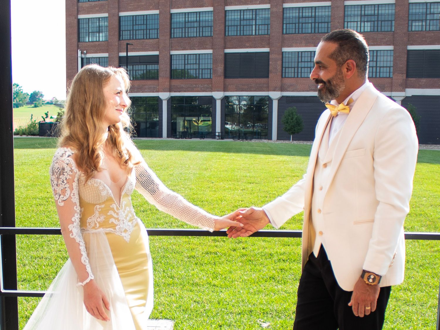 Couple in formal attire holding hands and walking together on a sunny green lawn outside a brick building.