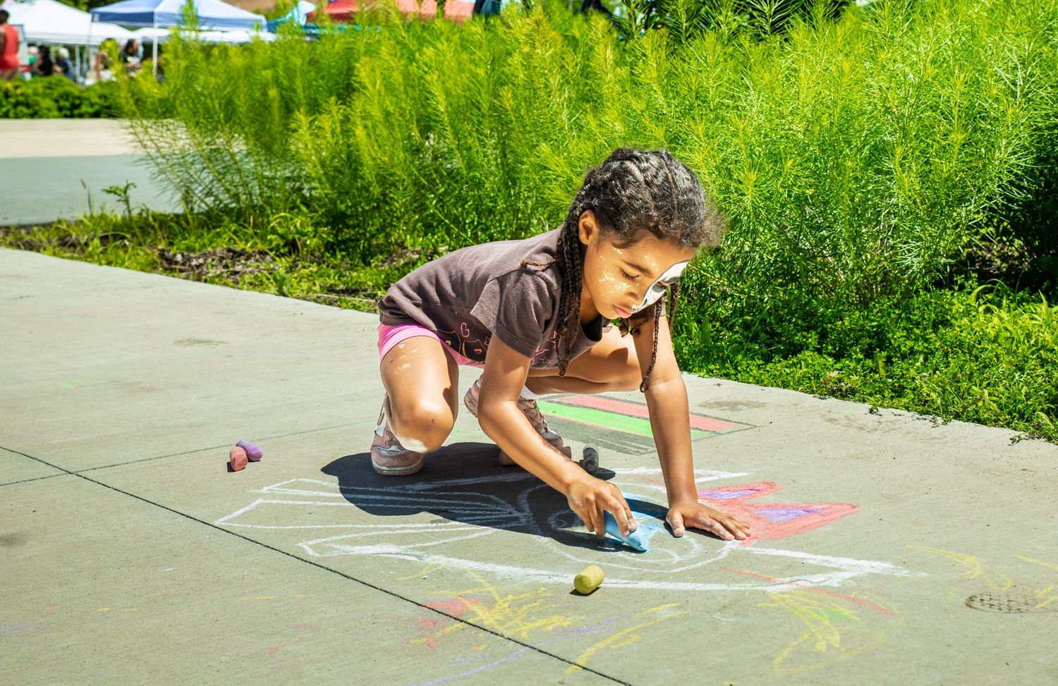 A young girl crouches on a sidewalk, drawing colorful chalk art outdoors on a sunny day.