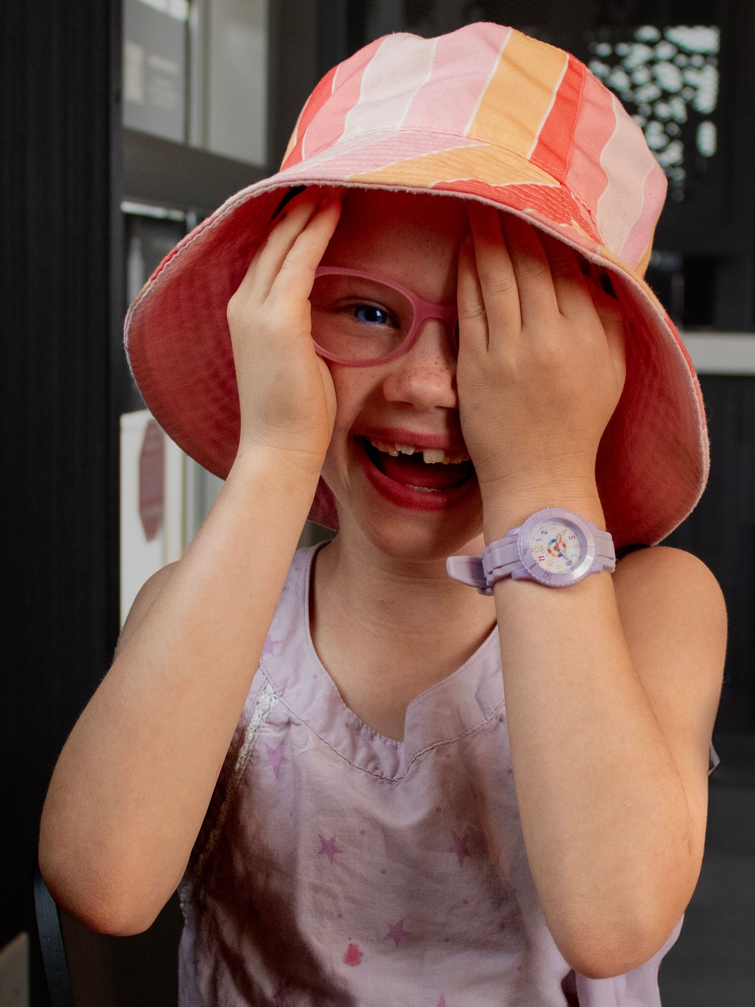 A young child in a pink sun hat plays peek-a-boo by covering their eyes while smiling and laughing.