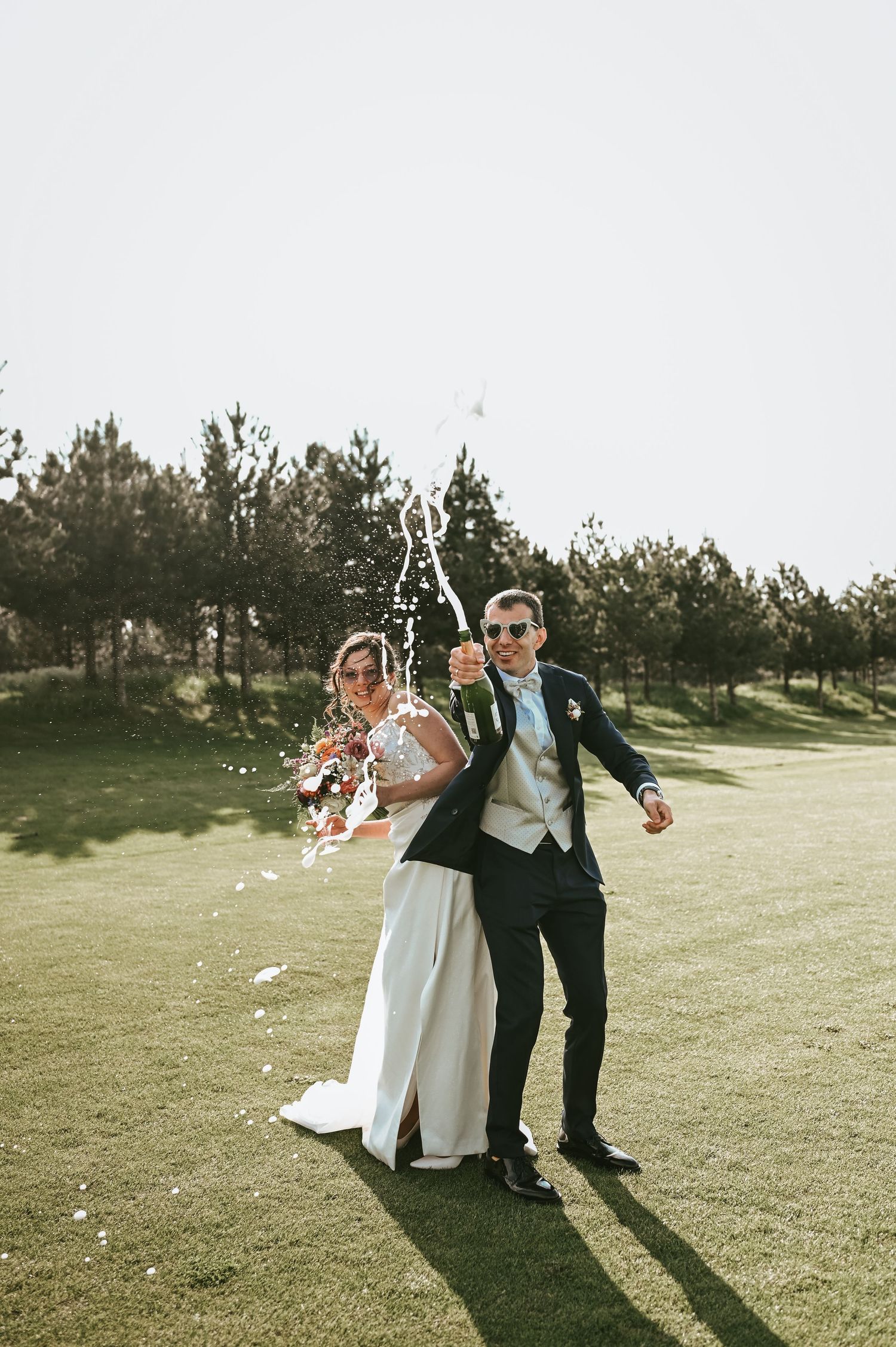 Wedding couple pops champagne and celebrates on a grassy hillside with pine trees in background.