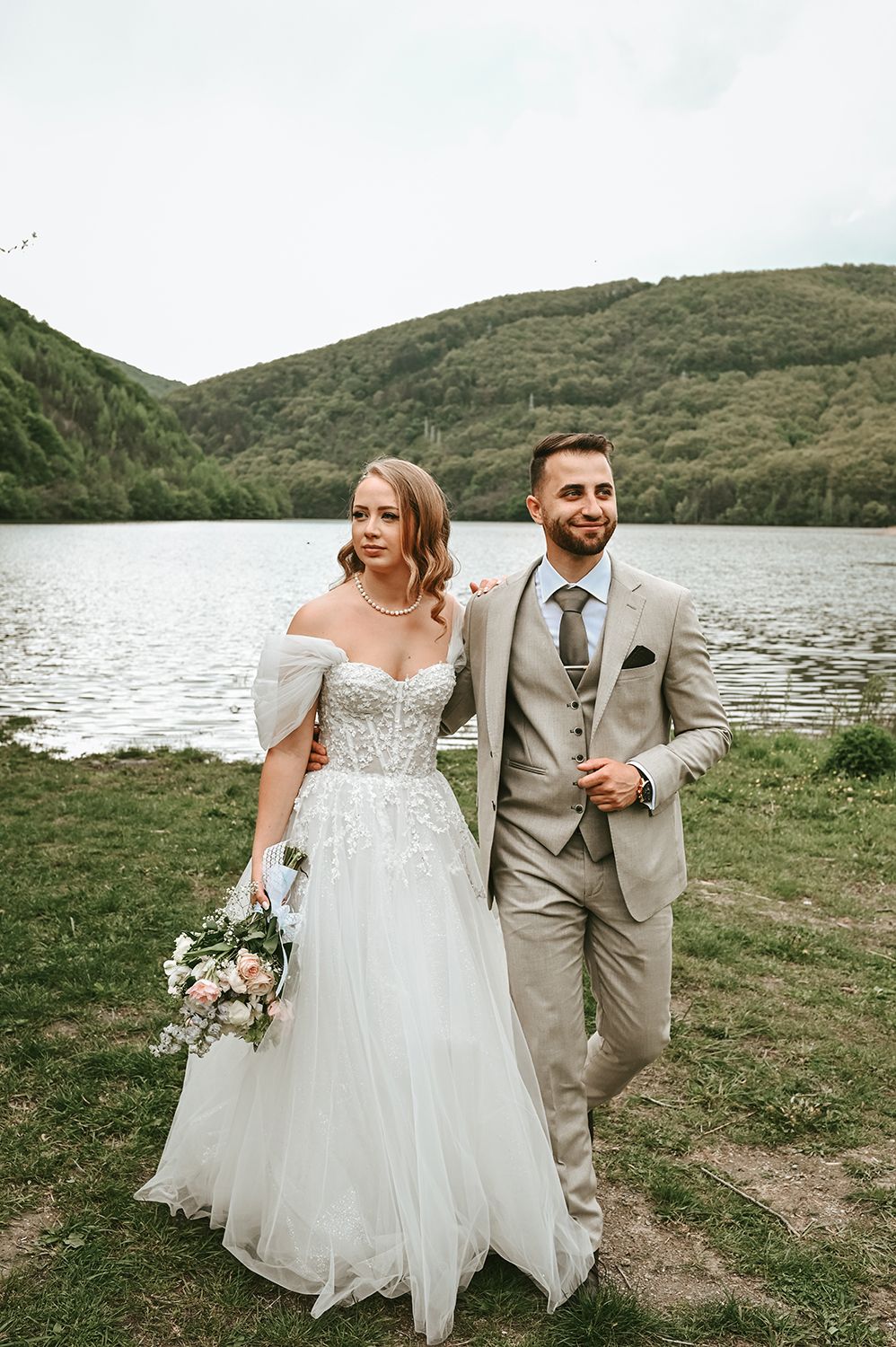 Elegant wedding couple poses beside a serene lake with rolling hills in the background.