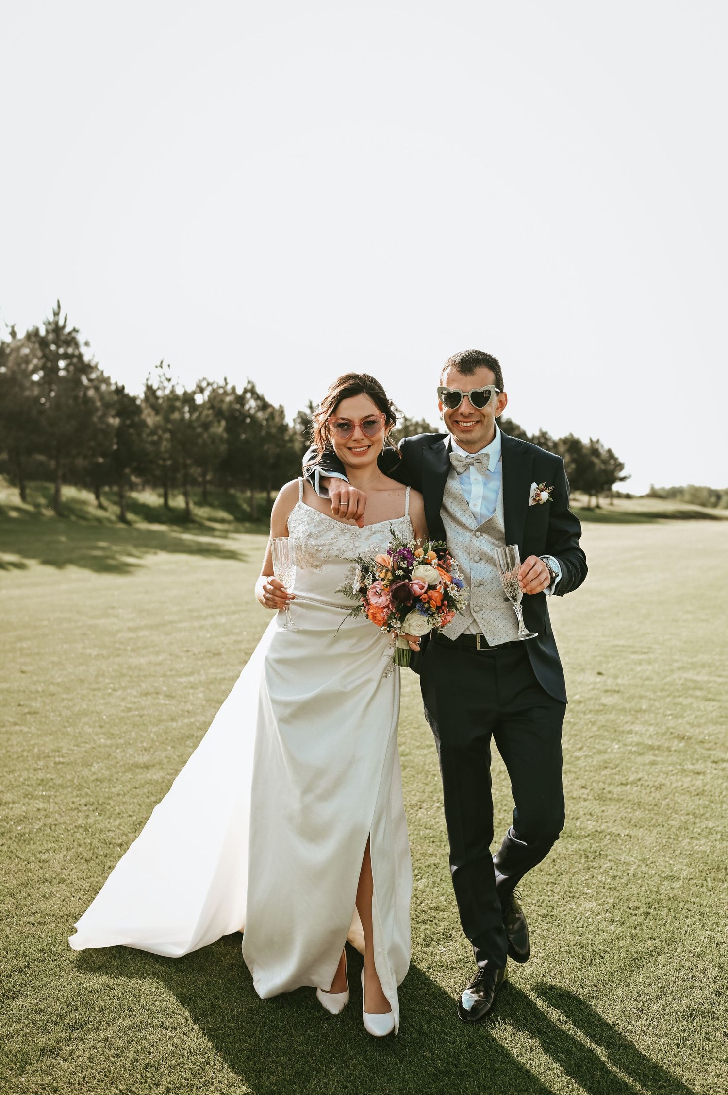 Stylish wedding couple in sunglasses walk across a golf course with a beautiful bouquet.