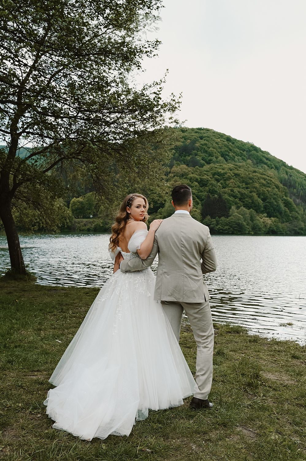 Wedding couple stands by a tranquil lake with lush green mountains creating a scenic backdrop.