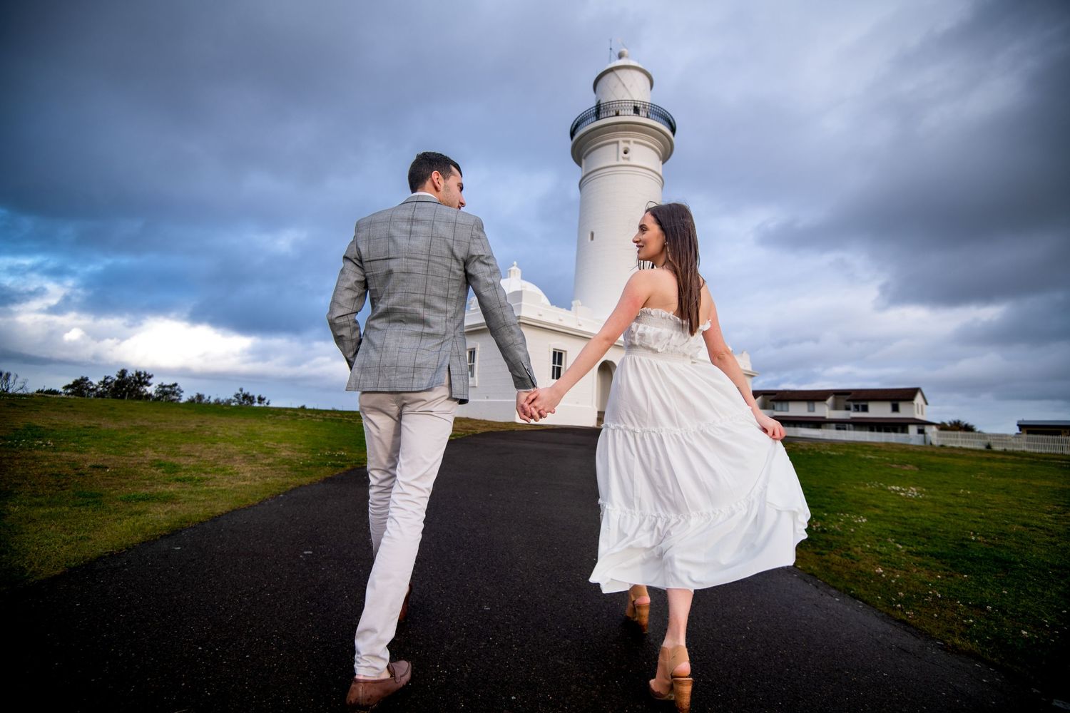 A couple in wedding attire holds hands while walking on a path toward a white lighthouse under stormy skies.