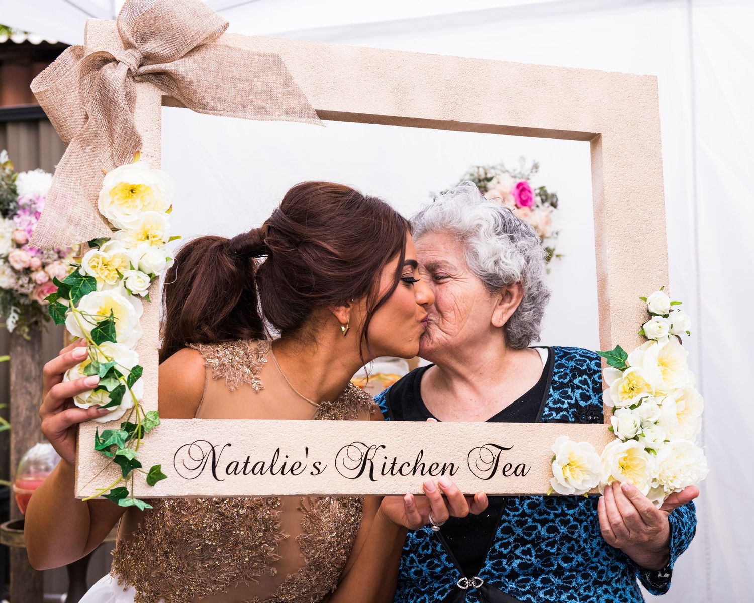Two people share an intimate kiss while surrounded by white and pink decorative floral arrangements.