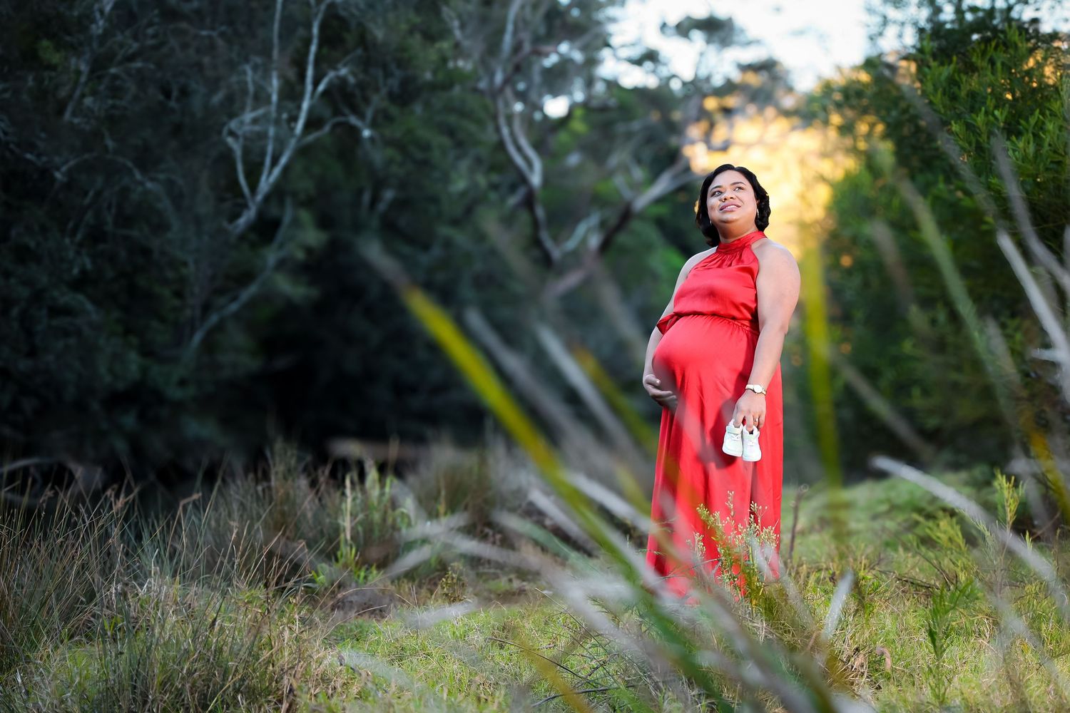 Figure in flowing red dress stands among trees at sunset.