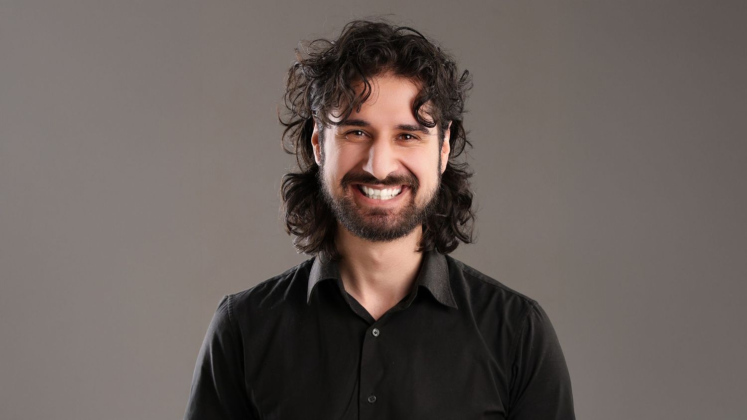 A person with dark curly hair and a beard wearing a black collared shirt stands against a grey backdrop. Professional corporate headshot.