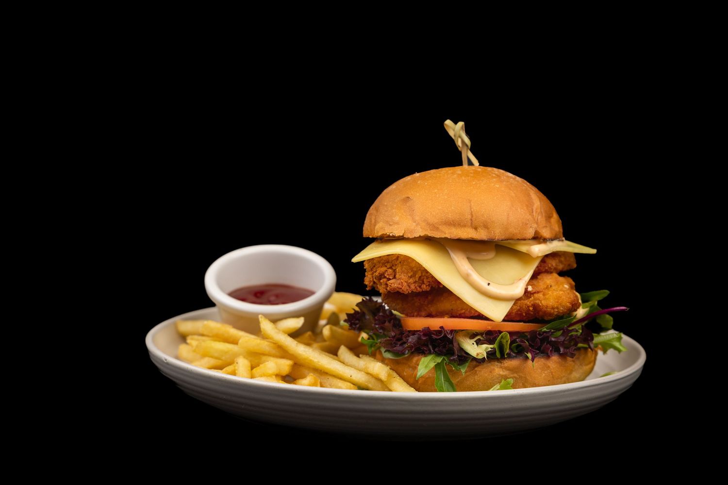 Juicy burger with melted cheese, crispy chicken, fresh veggies on a white plate with golden fries and ketchup on black background.