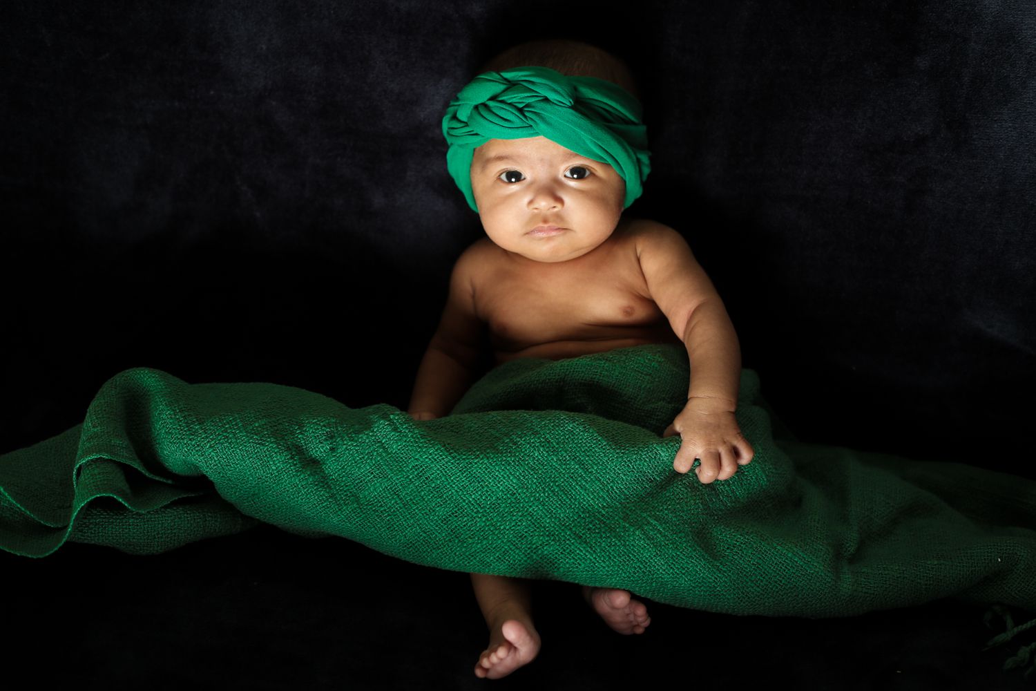 Alert baby wearing green headwrap on dark background with matching fabric.