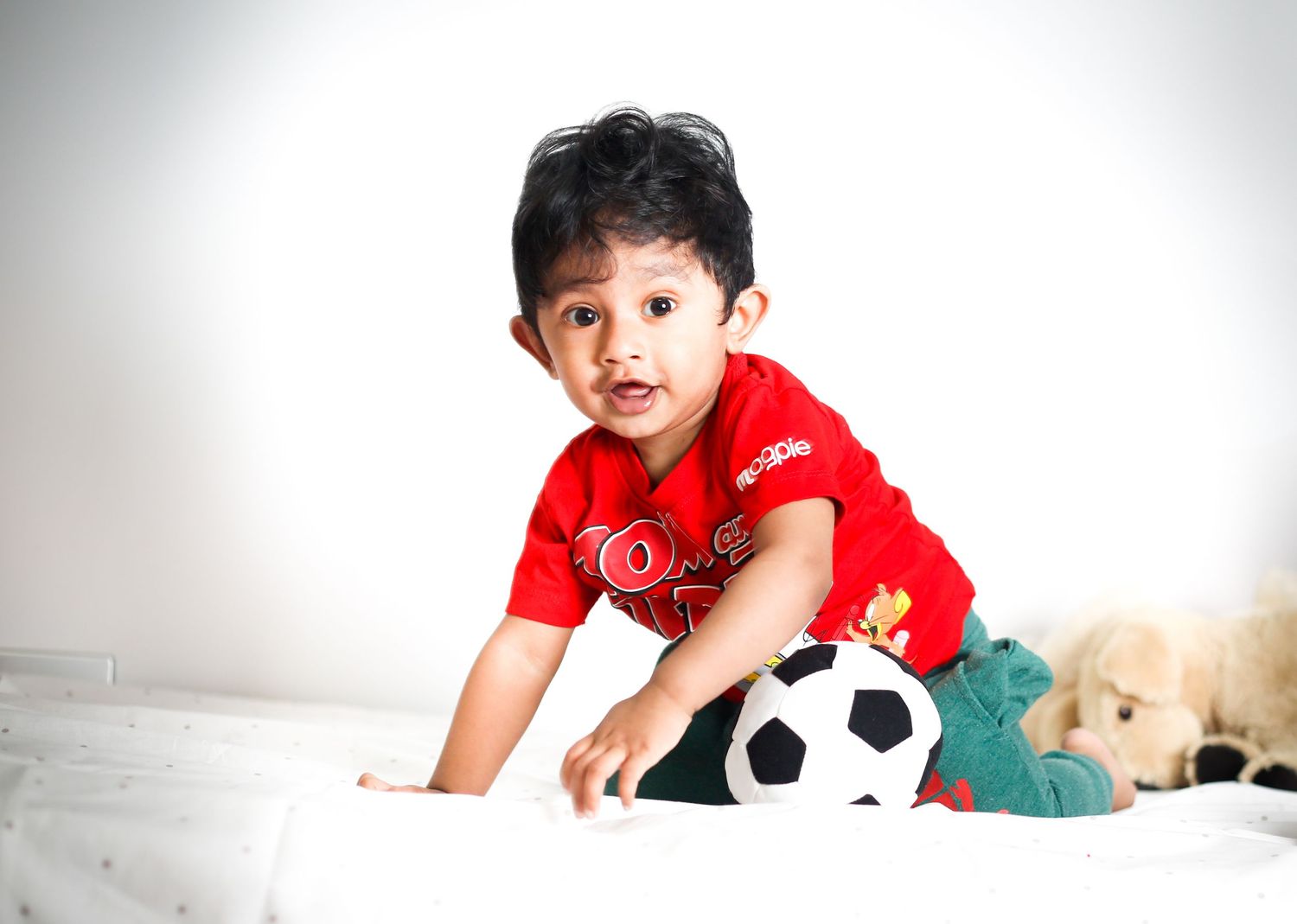 Young toddler in red t-shirt plays on floor with soccer ball and plush toy against white background.