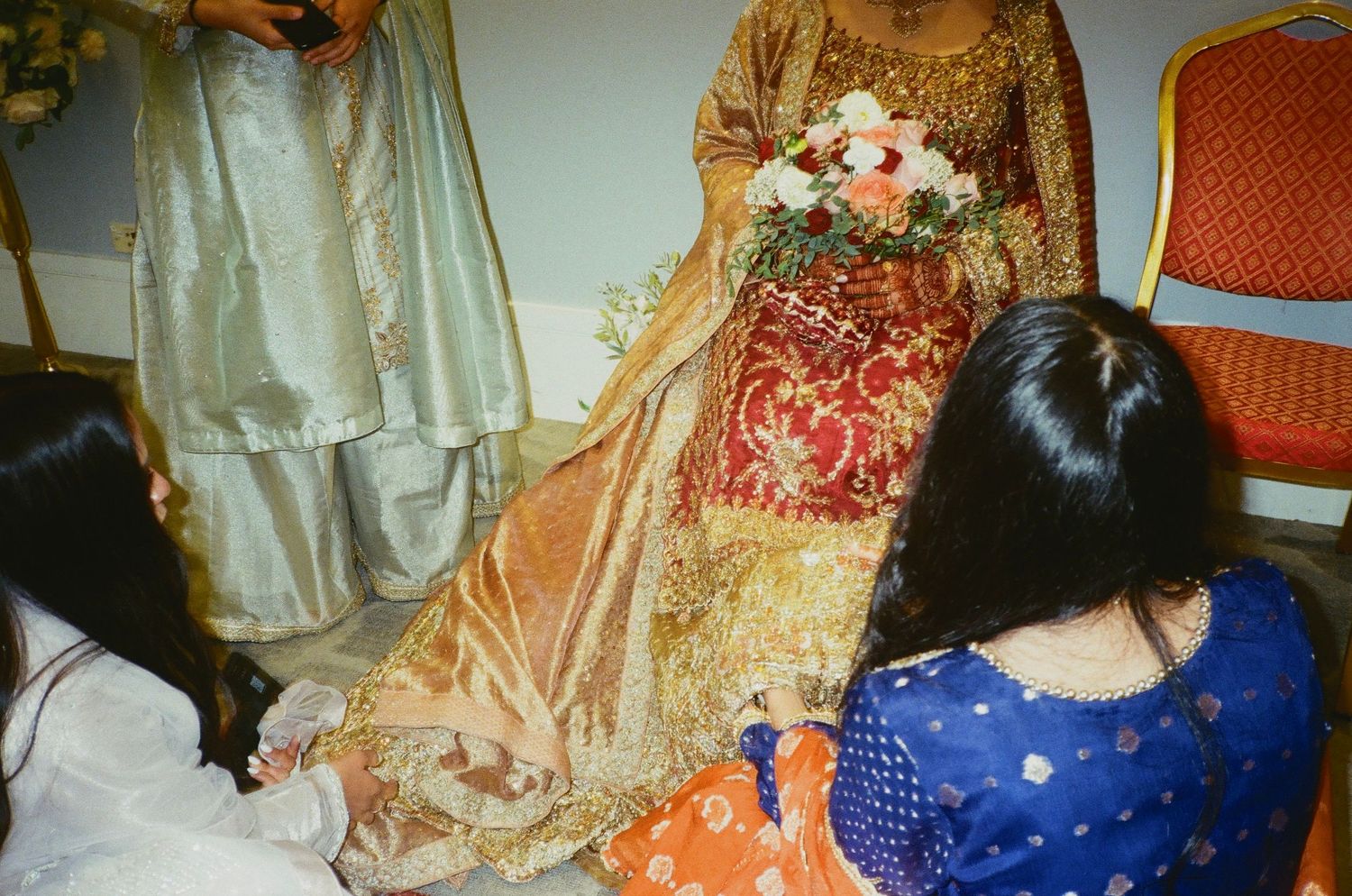 Bridesmaids help a red-dressed Desi bride adjust her outfit—surrounded by vibrant colors, captured on warm 35mm film in Chicago.south asian