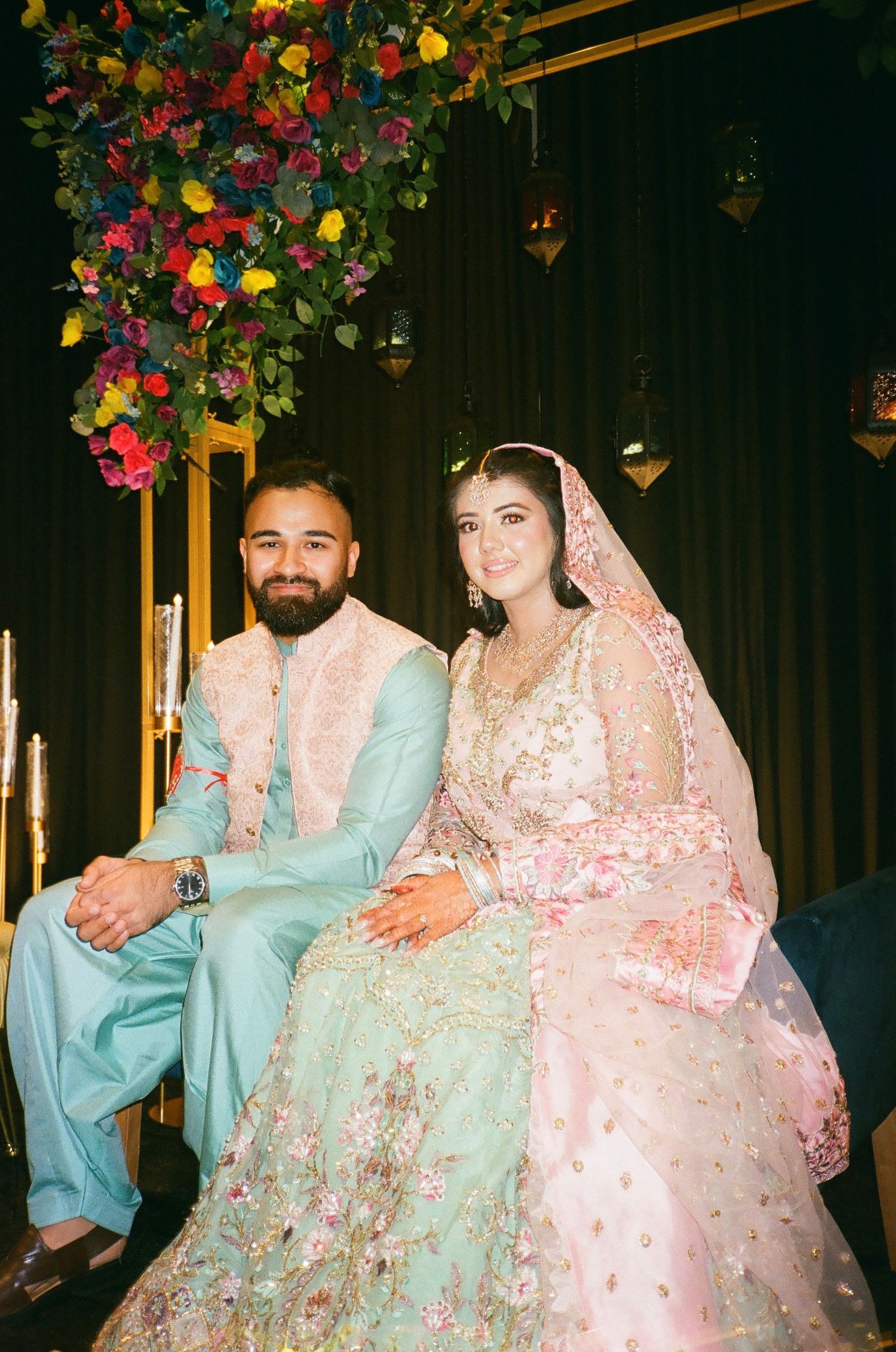 Mehndi-ready couple on stage in colorful pastels—playful, vibrant, and ready to celebrate, shot on vintage 35mm film. south asian