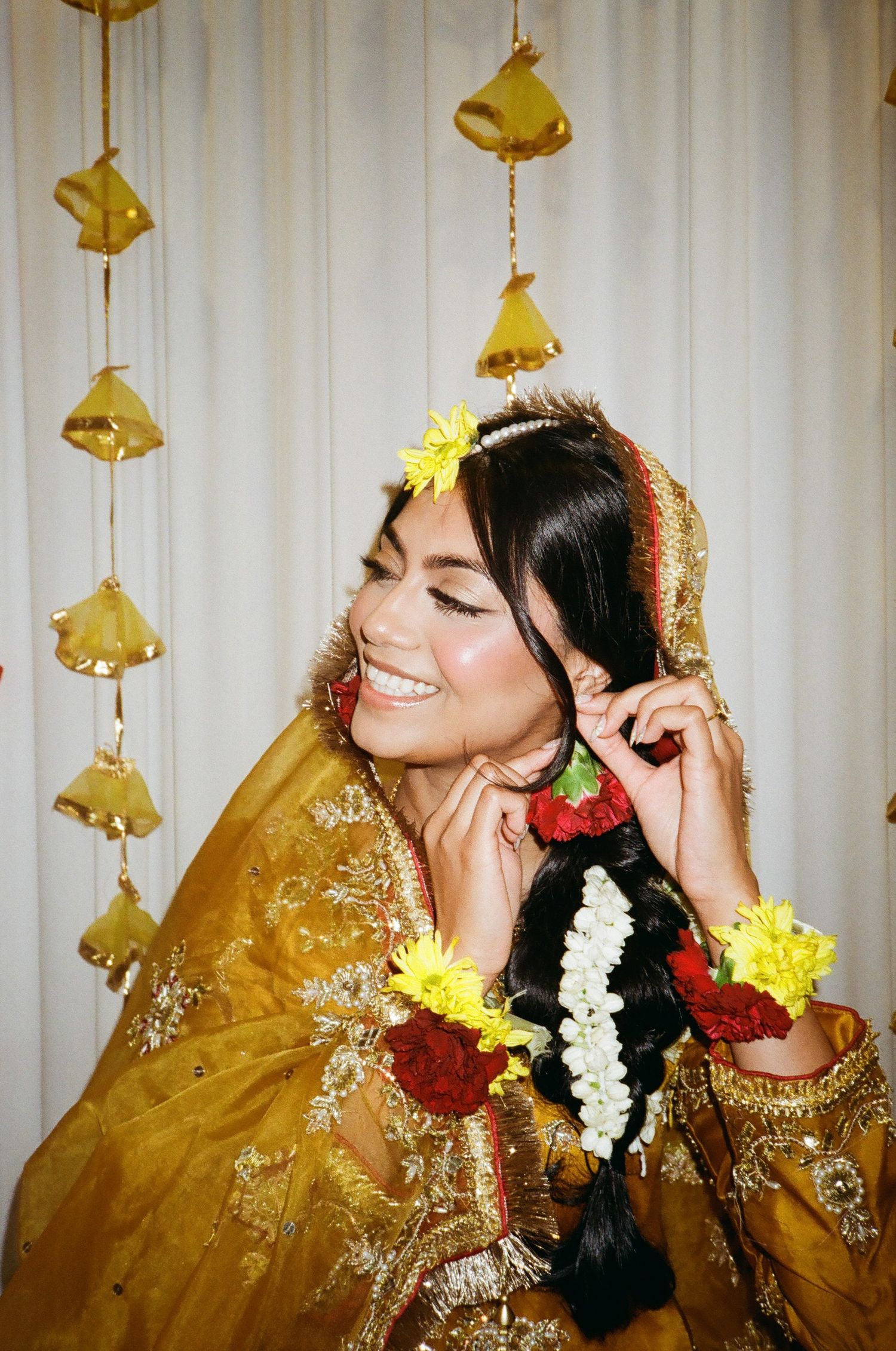 Desi Muslim bride at her Mayoun in a yellow Pakistani dress and gajra, fixing floral earrings—captured on 35mm film with vintage charm.