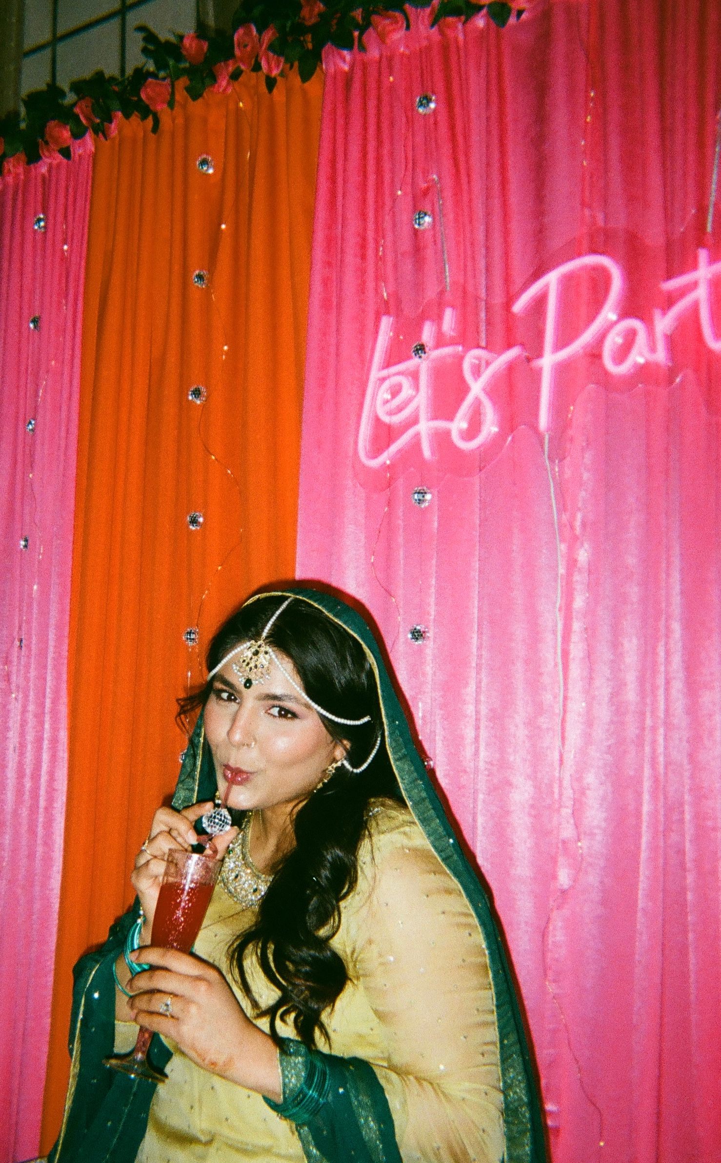 Bride-to-be relaxes, sipping a fun drink at her disco Dholki stage in Chicago suburbs, neon “Let’s Party” glowing—captured on 35mm film!