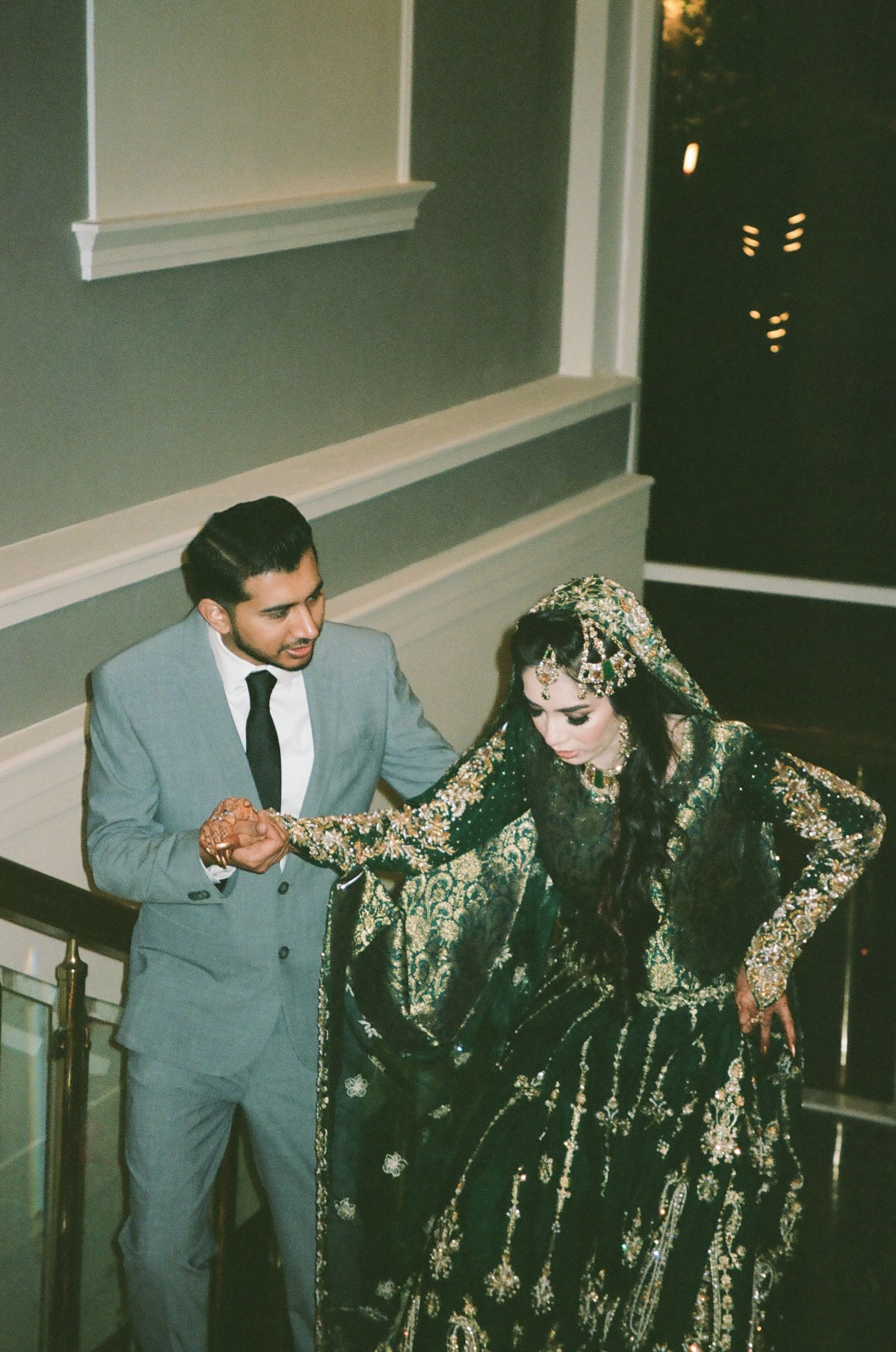Groom in grey gently helps his bride in emerald green ascend the stairs—royal Desi vibes, cinematic light, and 35mm timelessness.