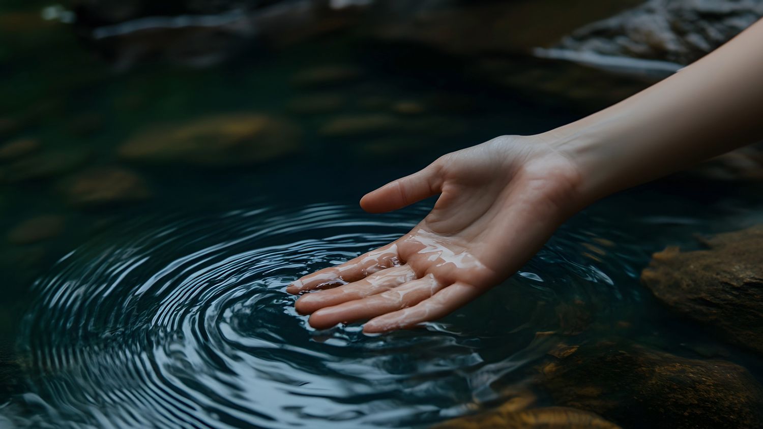 A hand gently touches the surface of dark water, creating rippling circles.