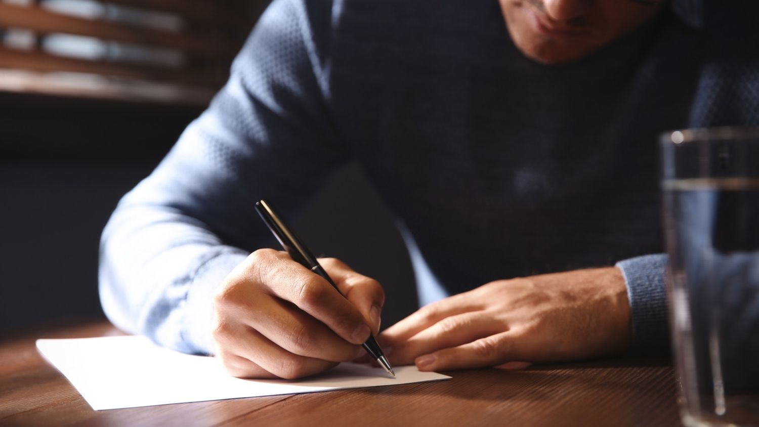 Someone writing on paper with a pen while seated at a desk near a window.