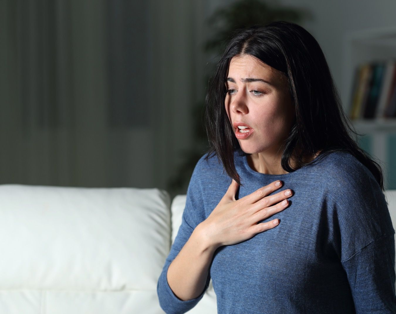 Woman in blue shirt clutches chest with distressed expression while sitting on white couch in dim room.