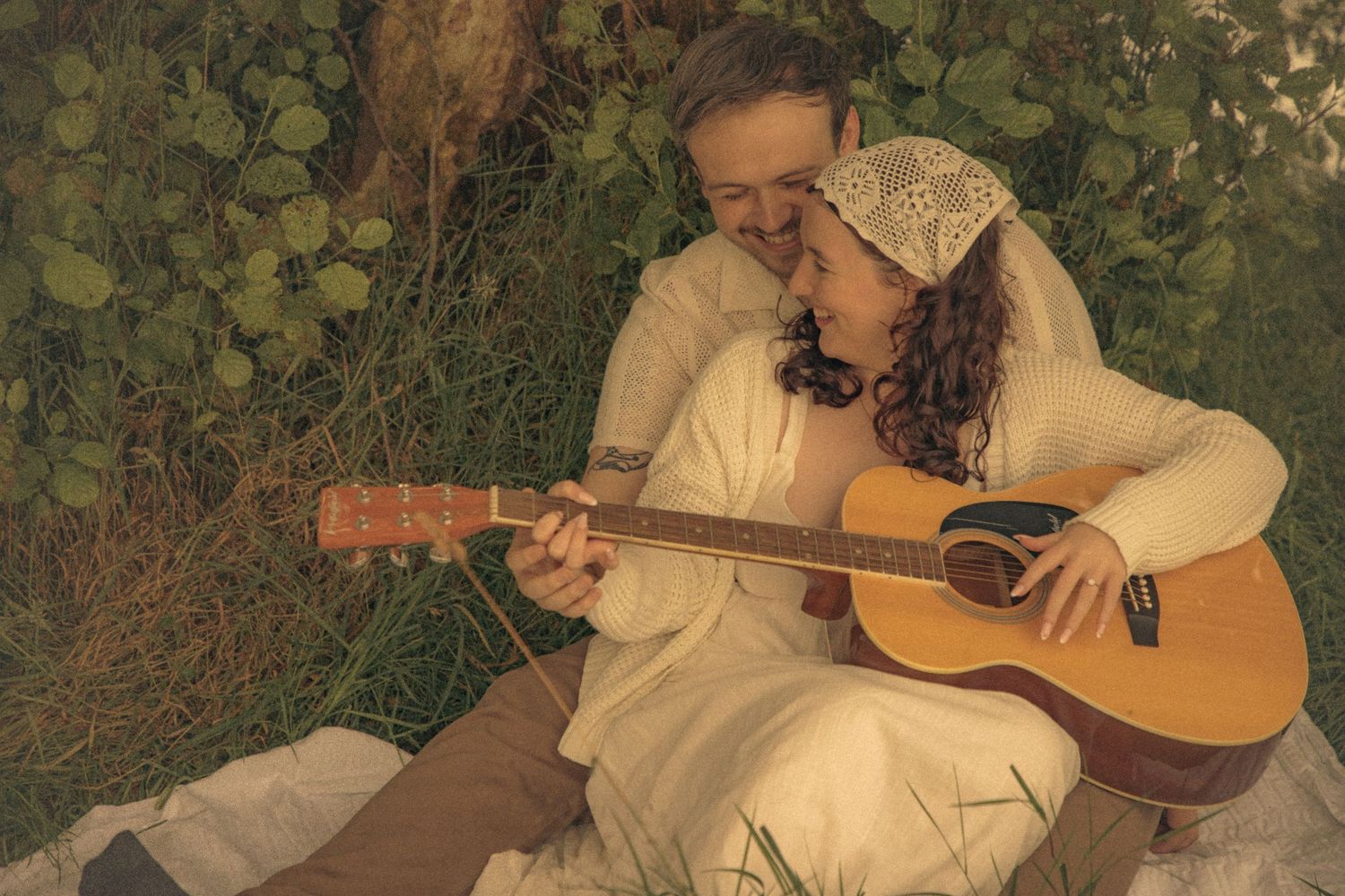 couple-sitting-on-the-grass-by-a-river-playing-guitar-together-during-a-cottagecore-style-engagement-photoshoot