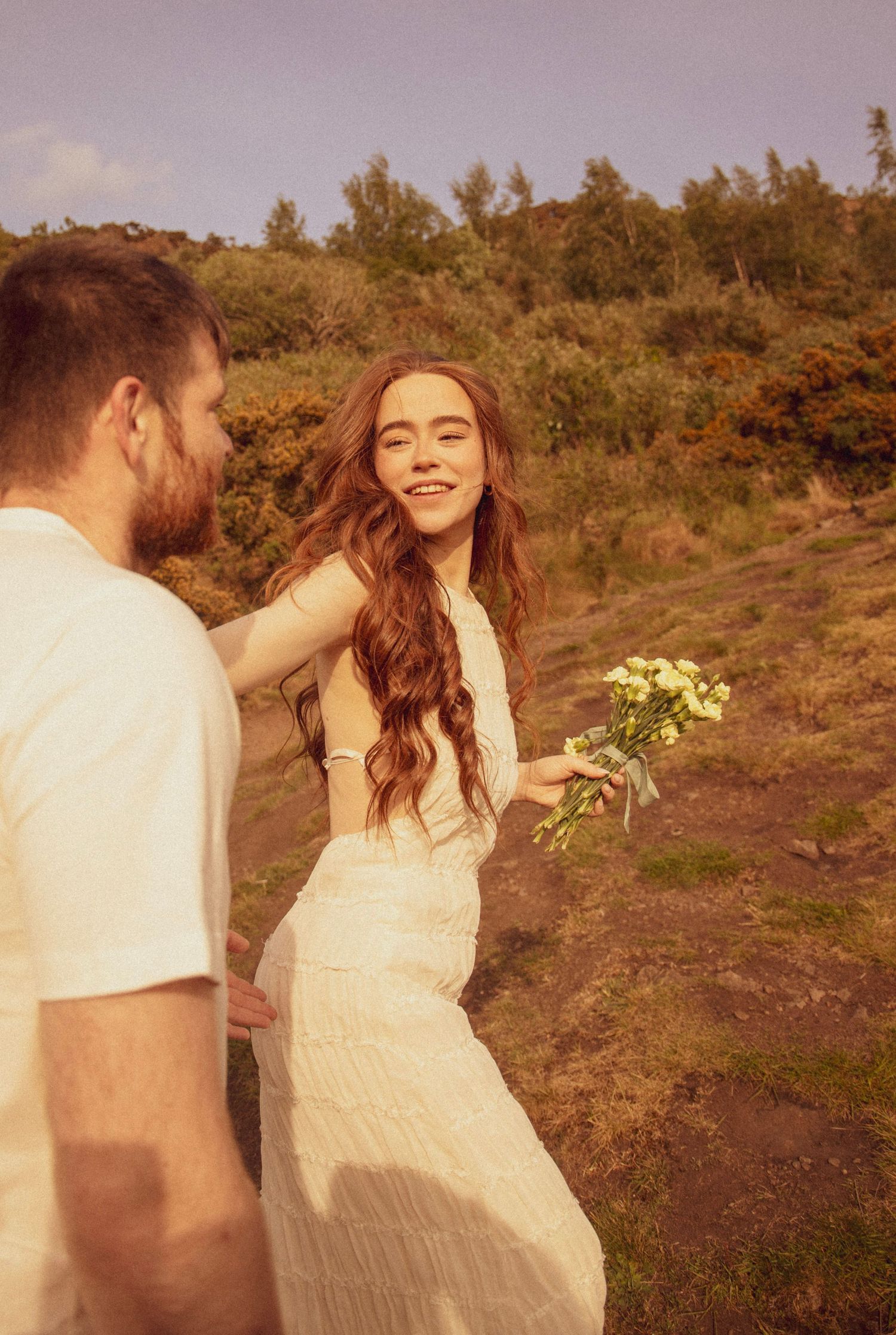 woman-holding-flowers-leading-boyfriend-up-a-hill-in-holyrood-park-edinburgh