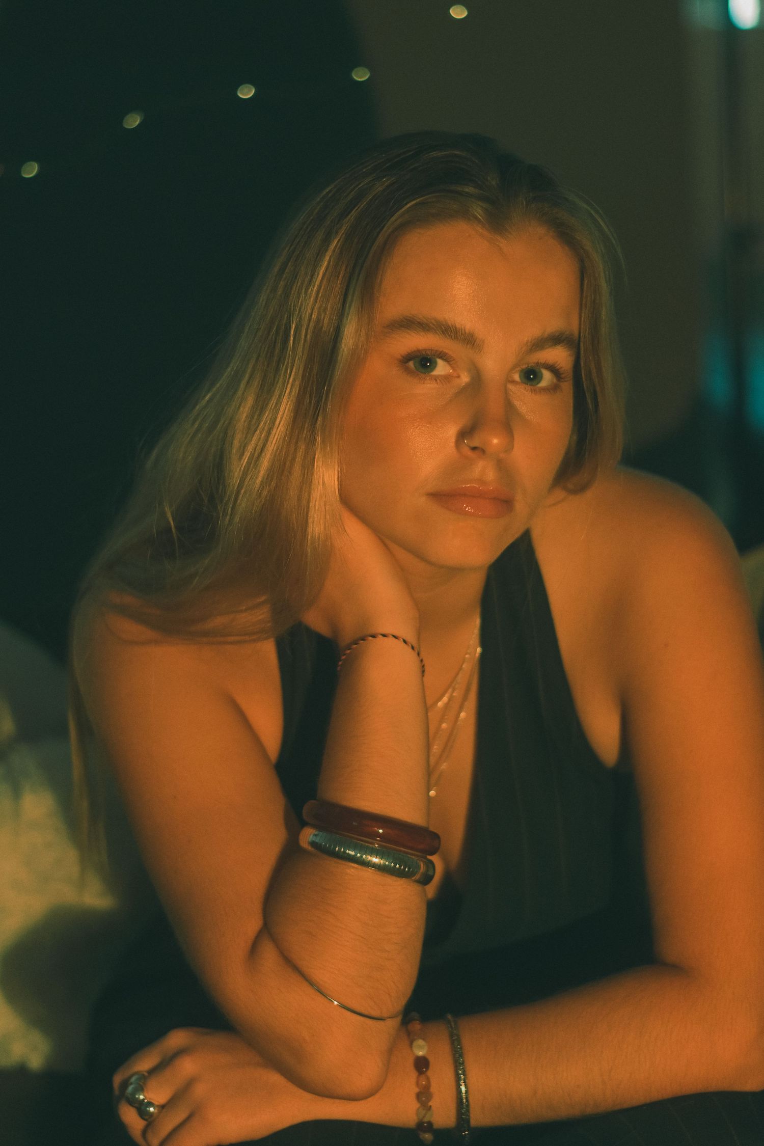 Moody portrait of a person in warm lighting wearing jewelry and a dark top.