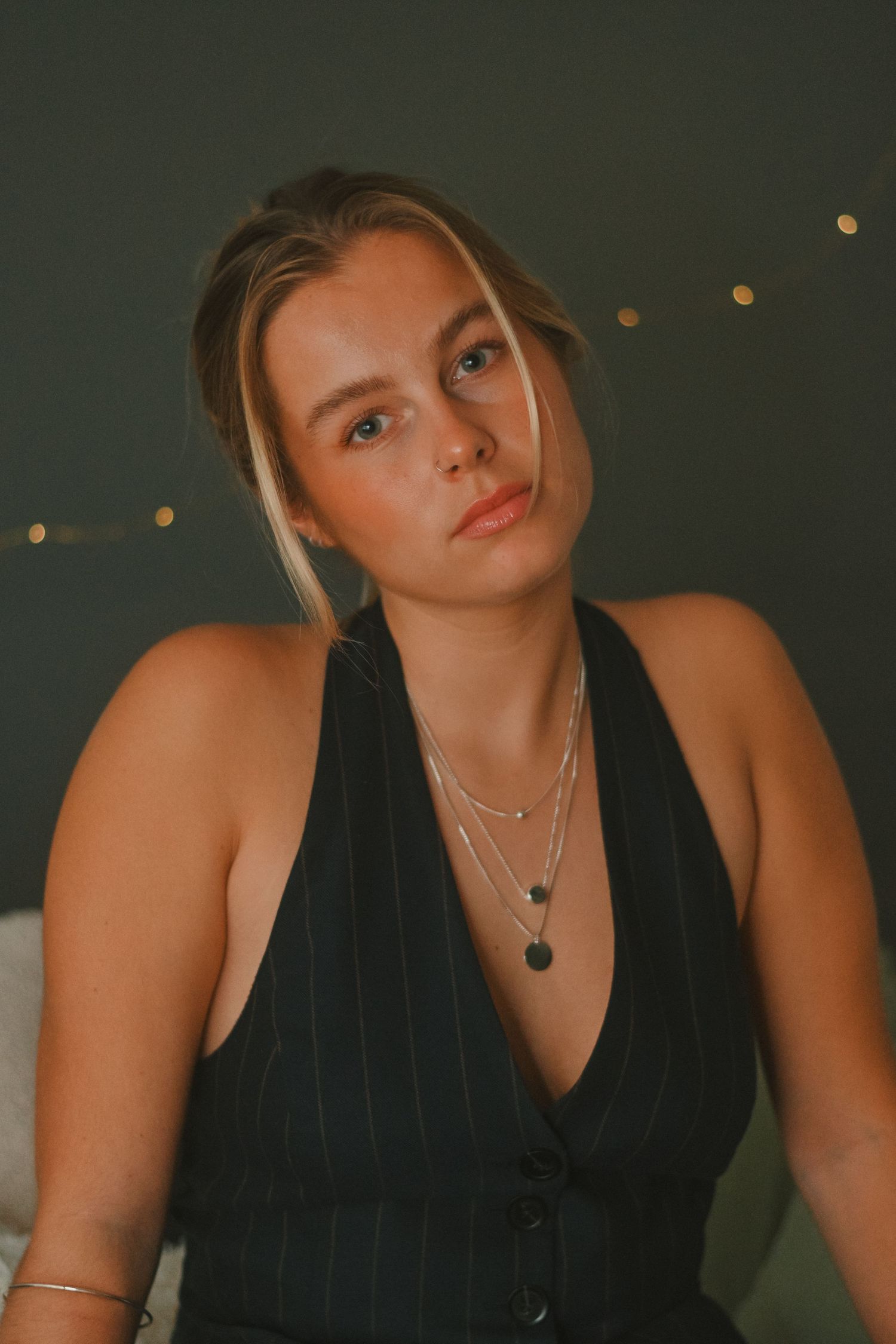 Elegant portrait in pinstripe vest with layered necklaces against dark background.