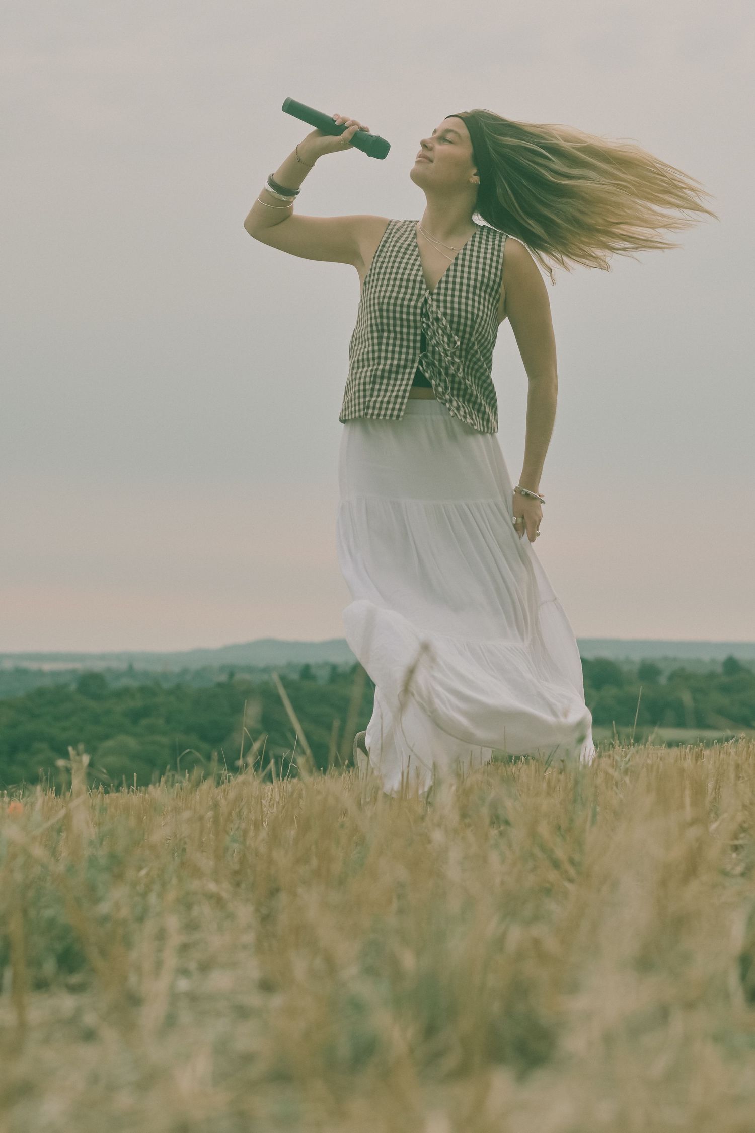 Person twirls in flowy white dress through tall grass with mountains in distance.