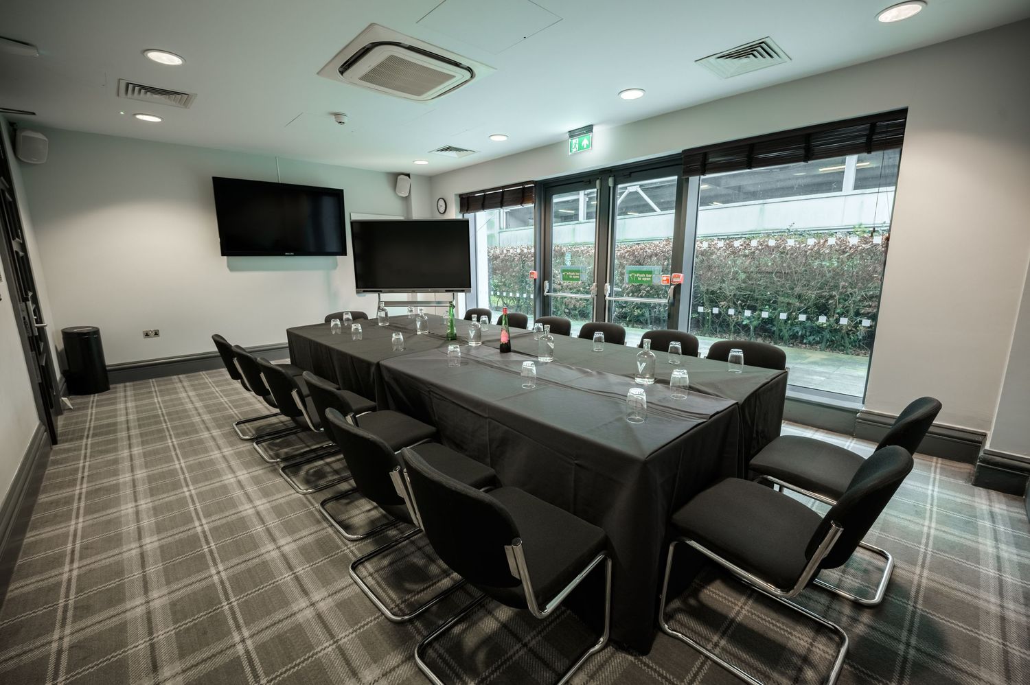 Modern boardroom with long black conference table, ergonomic chairs, wall-mounted TV, and large windows overlooking greenery.