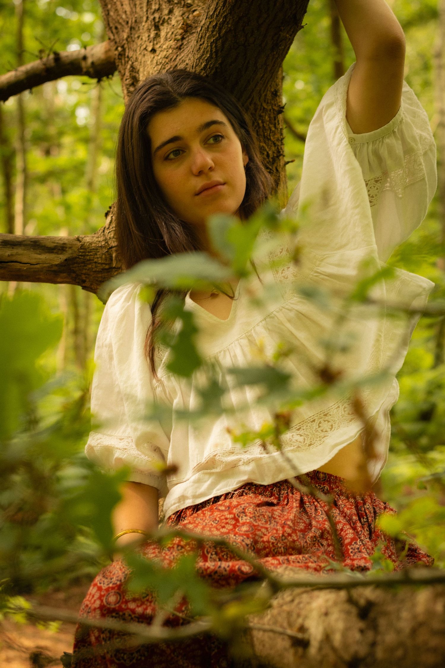 Person in white top and floral skirt sits dreamily in a sunlit forest setting among green foliage.