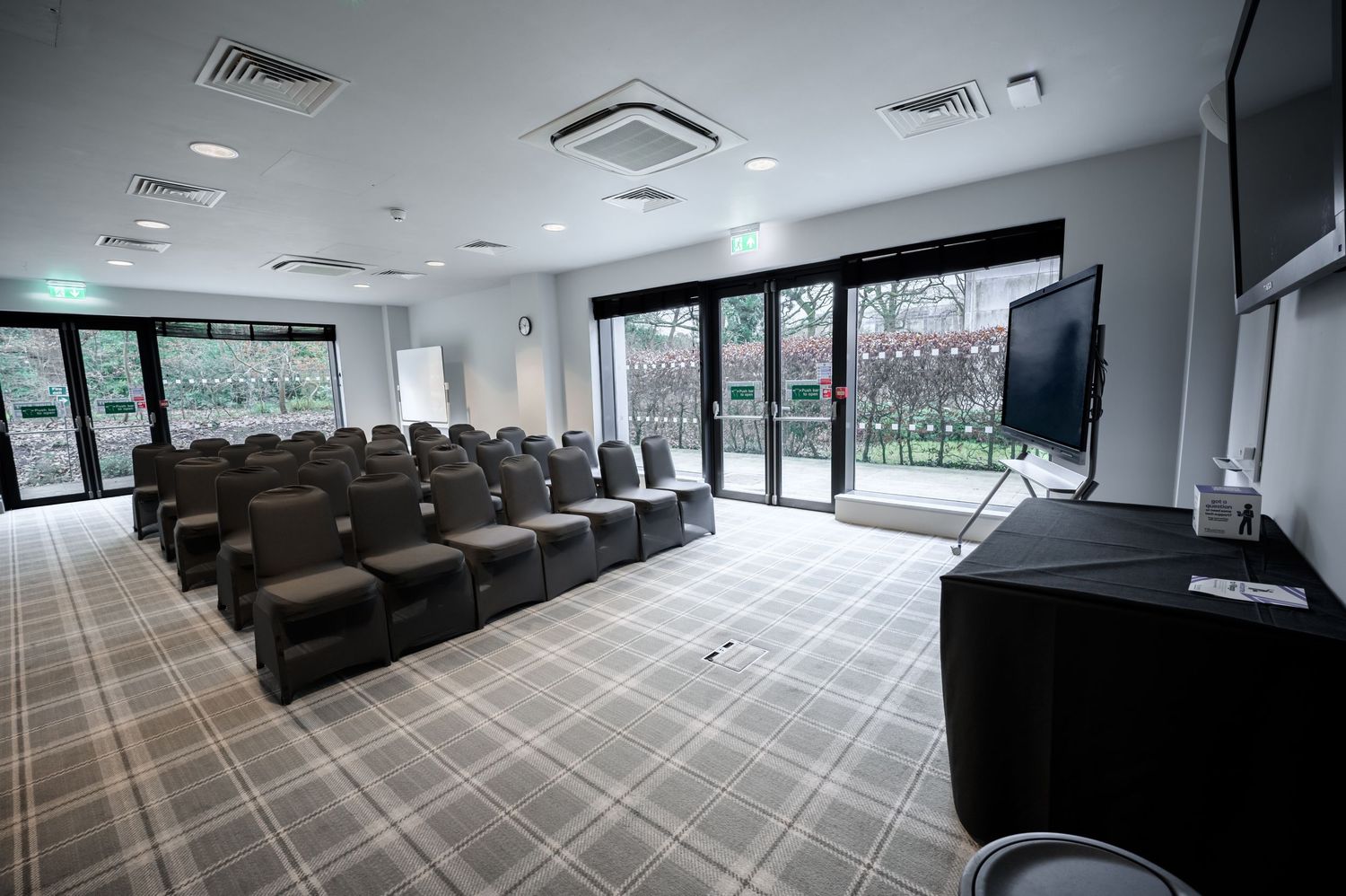 Modern conference room with rows of dark chairs, large windows, patterned carpet, and a presentation screen on a stand.