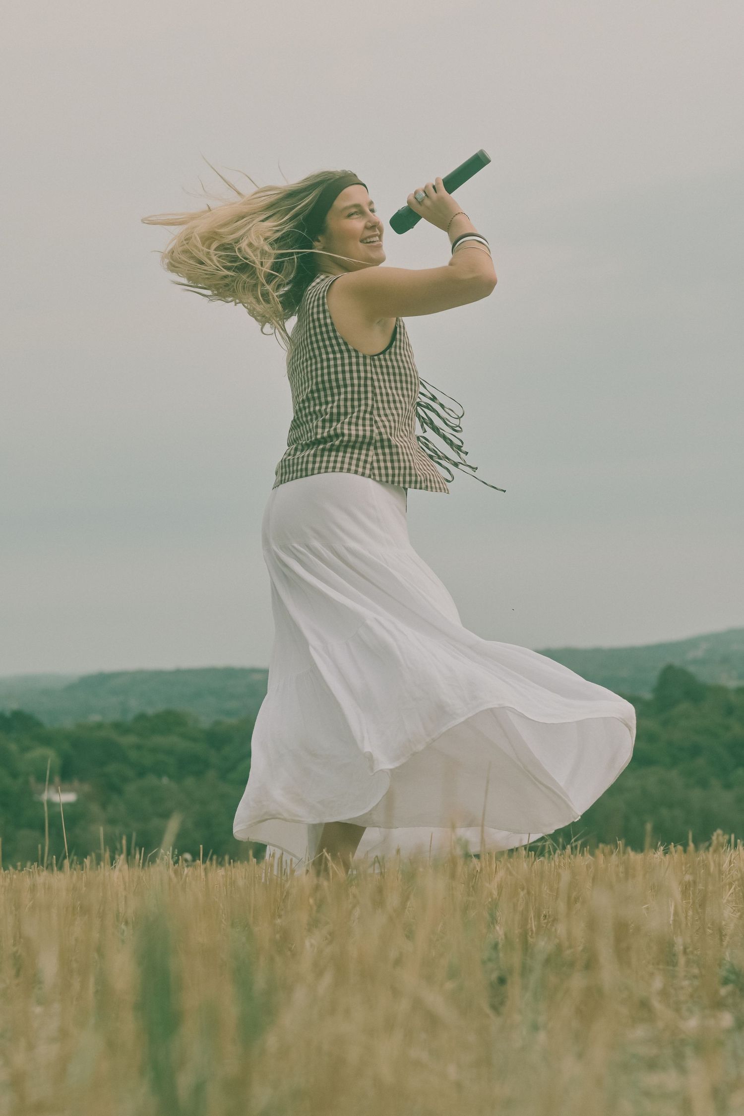 Person in white dress dances freely in sunlit field with mountains in background.