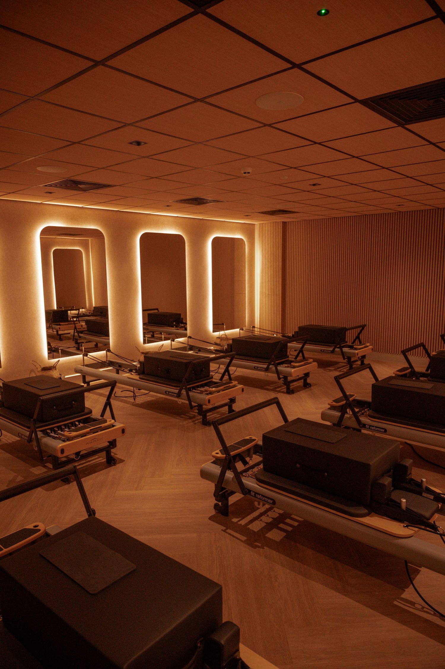 Warmly lit Pilates reformer studio with arched mirrors, ambient lighting, and multiple reformer machines on herringbone floors.