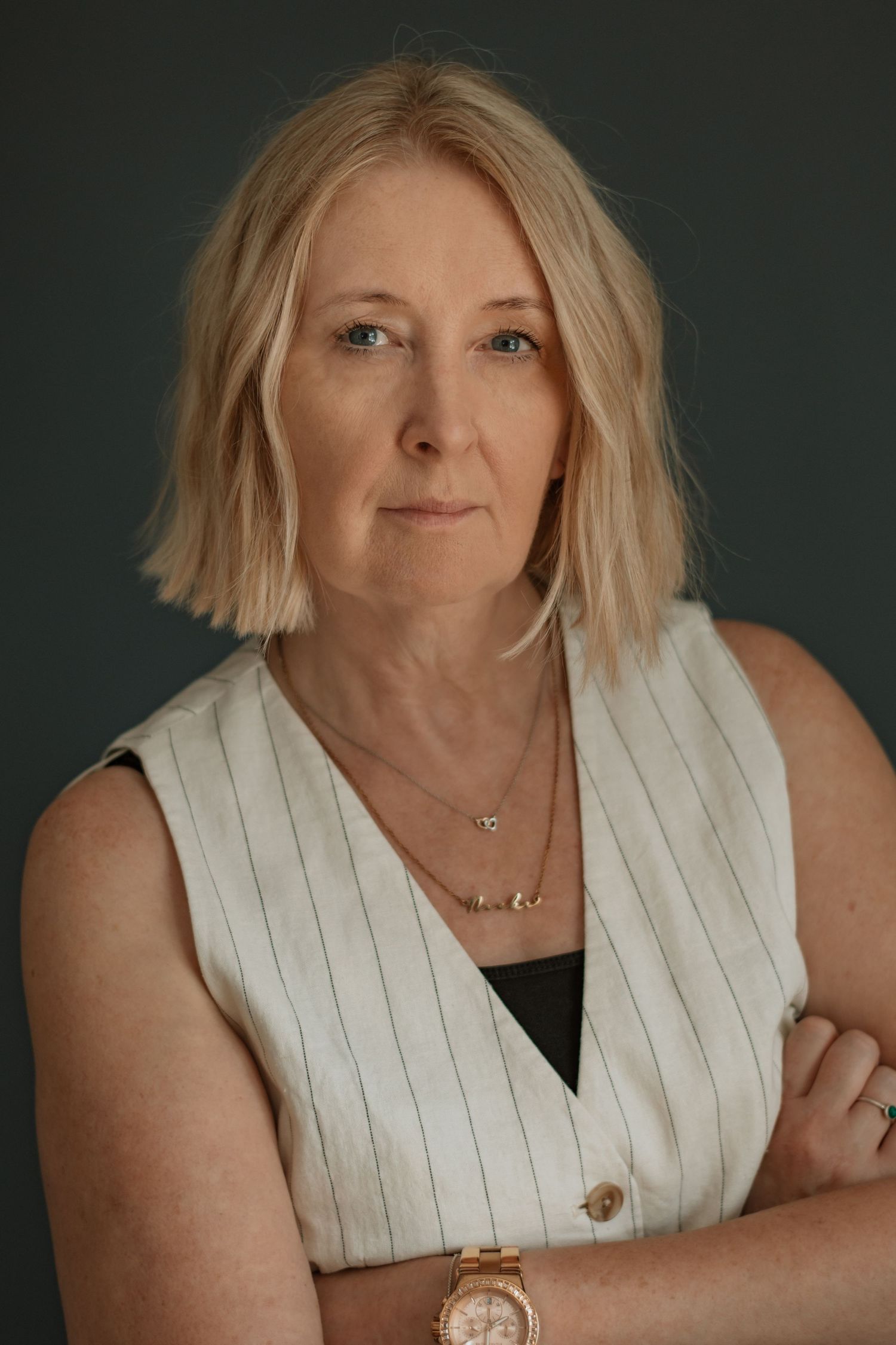Professional studio portrait against dark green background wearing white pinstriped vest and delicate necklace.