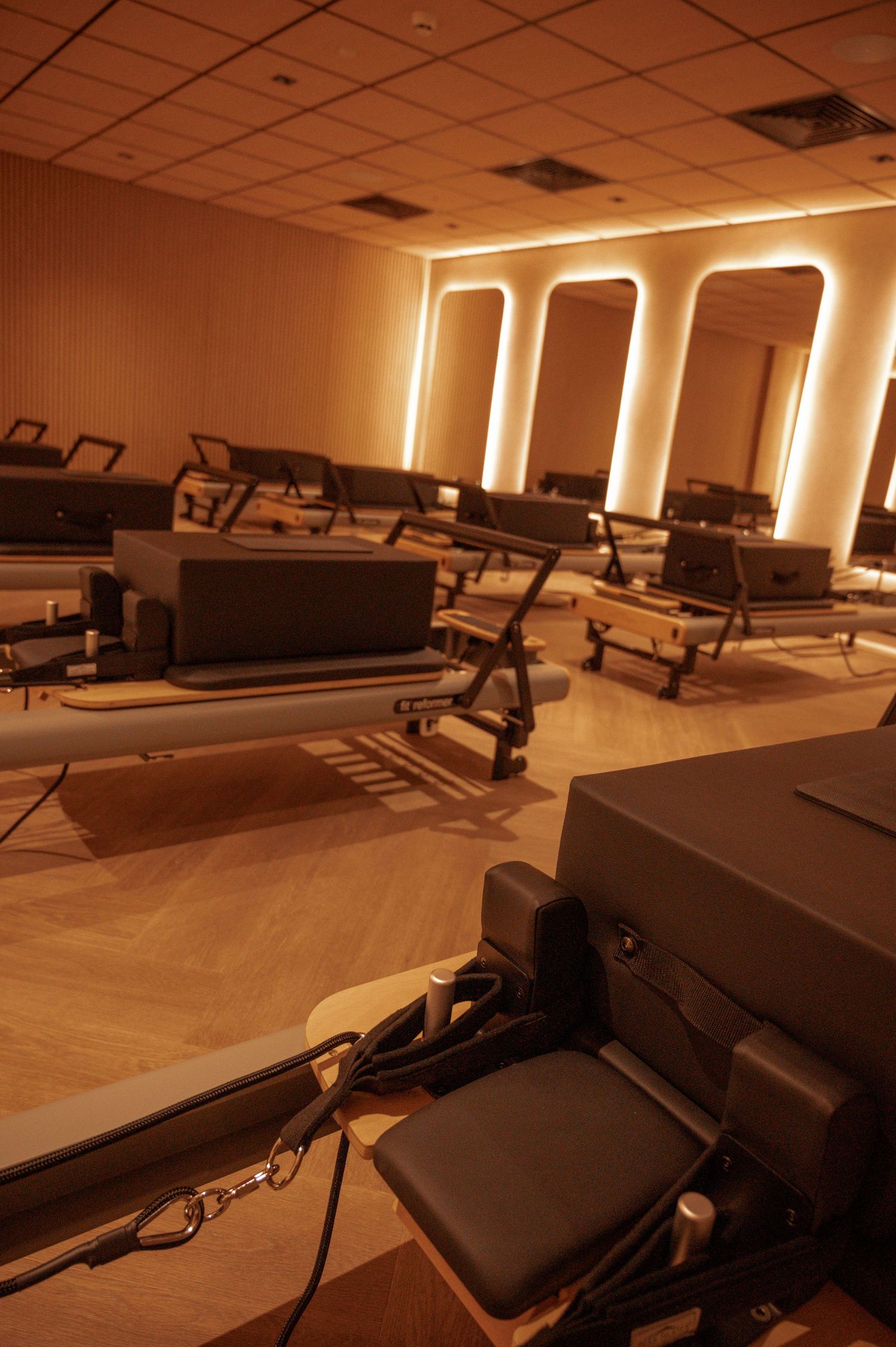 Empty Pilates reformer studio with warm golden lighting, arched mirrors, and wooden herringbone floors.