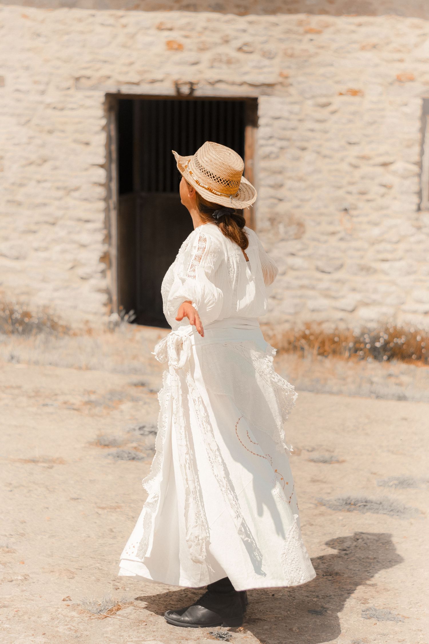 A figure in a flowing white vintage dress and straw hat twirls gracefully against a rustic stone wall background.