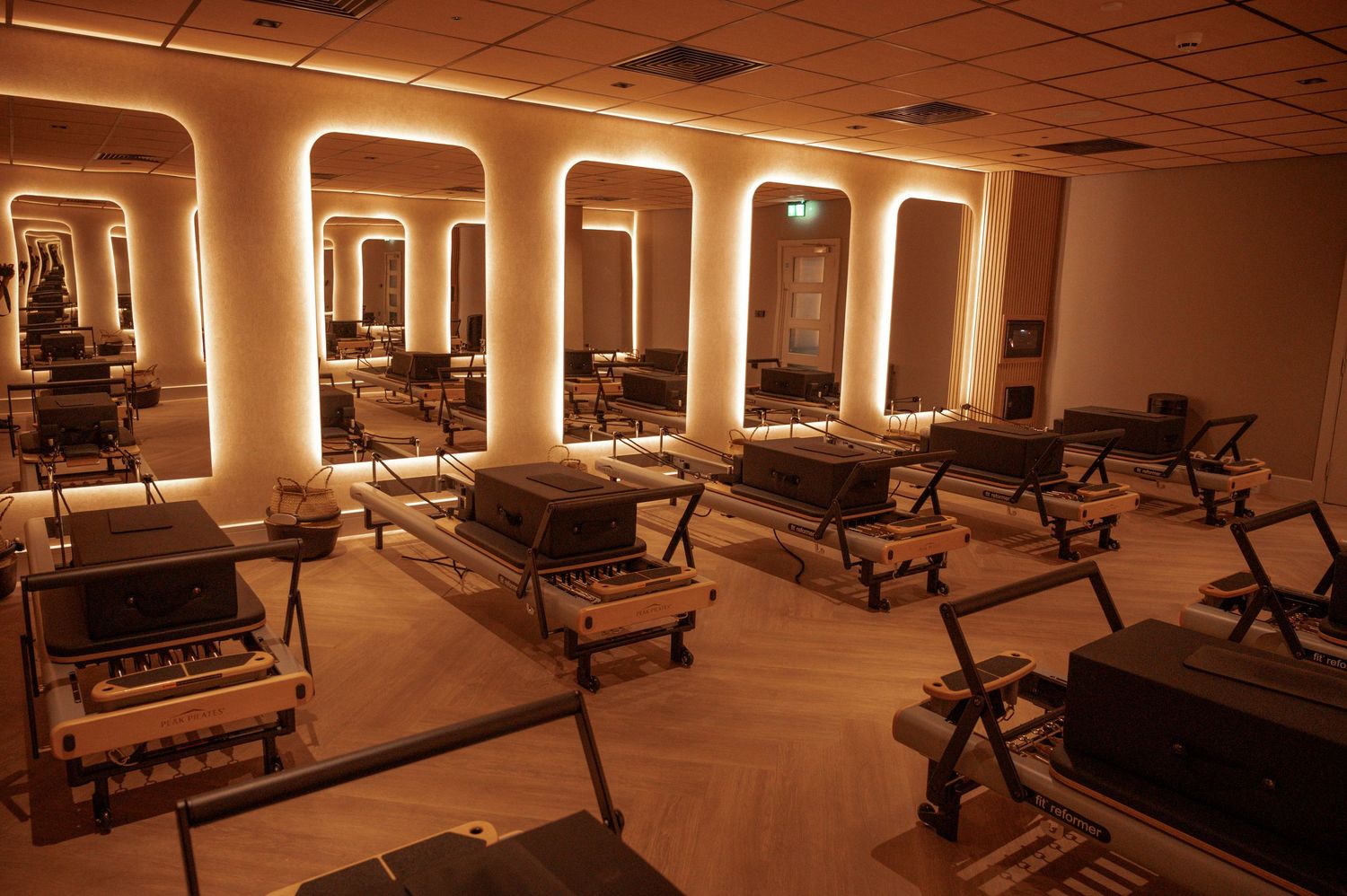 Luxury Pilates studio with reformer machines, arched mirrors, warm amber lighting, and herringbone wood floors.