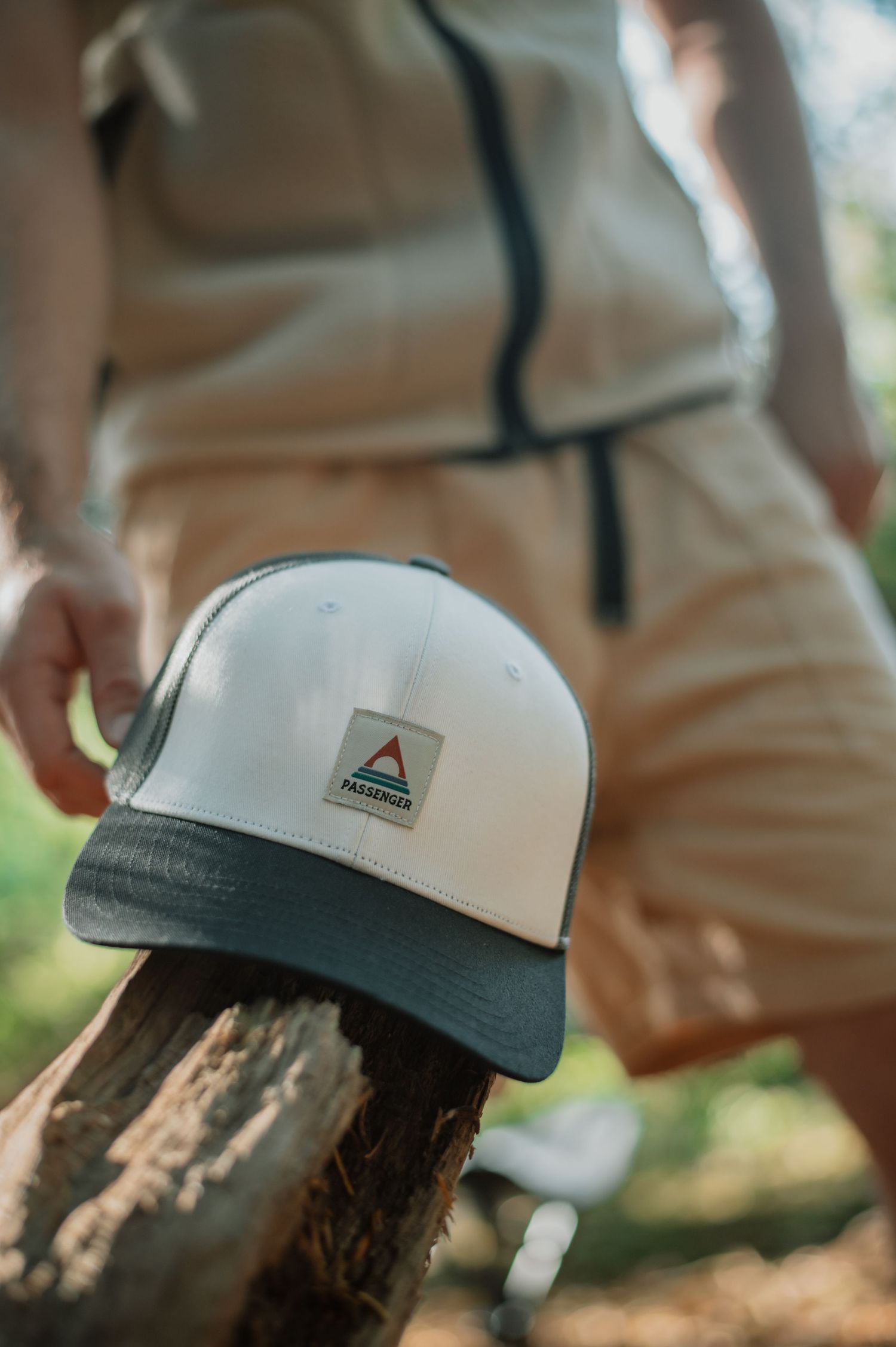 White and dark brim Passenger snapback cap resting on a log outdoors with a person in beige outfit behind it.