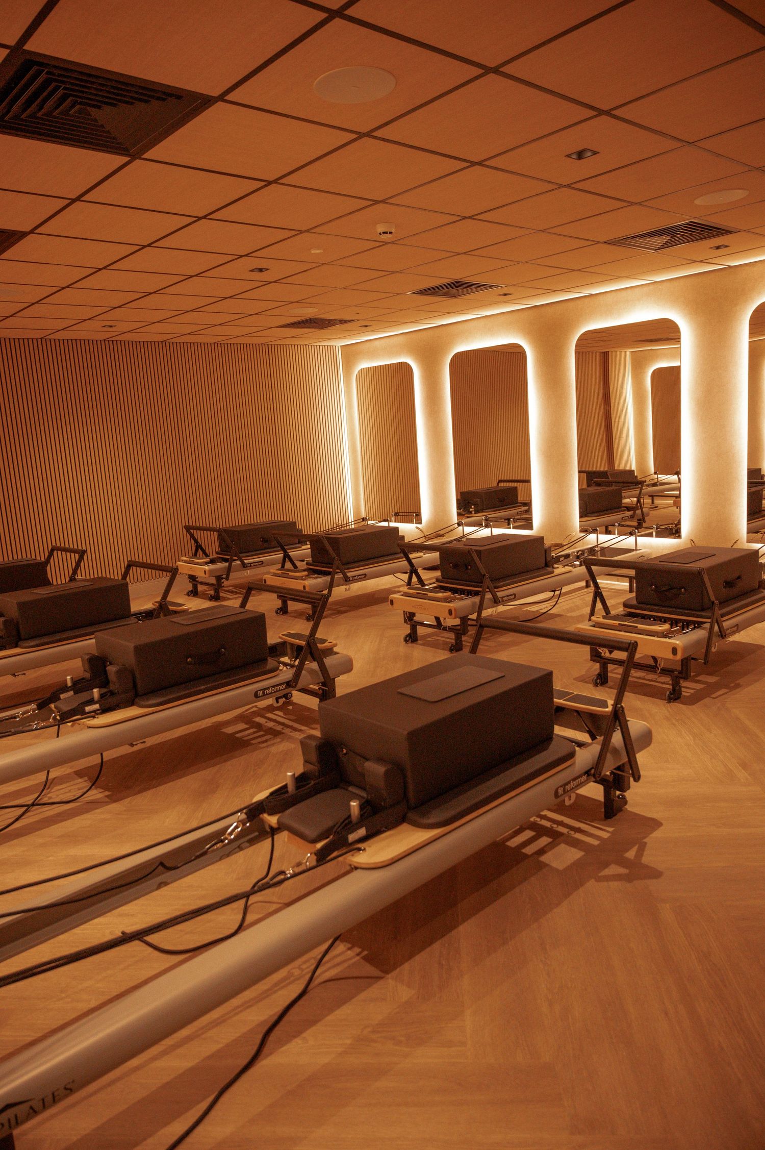 A warmly lit Pilates studio with reformer machines arranged in rows on hardwood floors, featuring arched mirror accents.