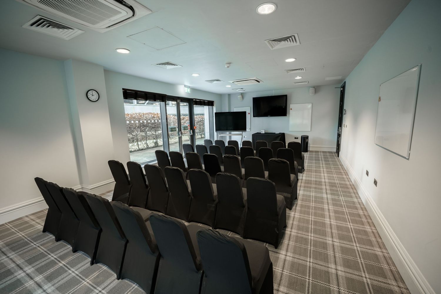 Modern conference room with rows of black chairs, wall-mounted TV, whiteboard, and large windows with natural light.