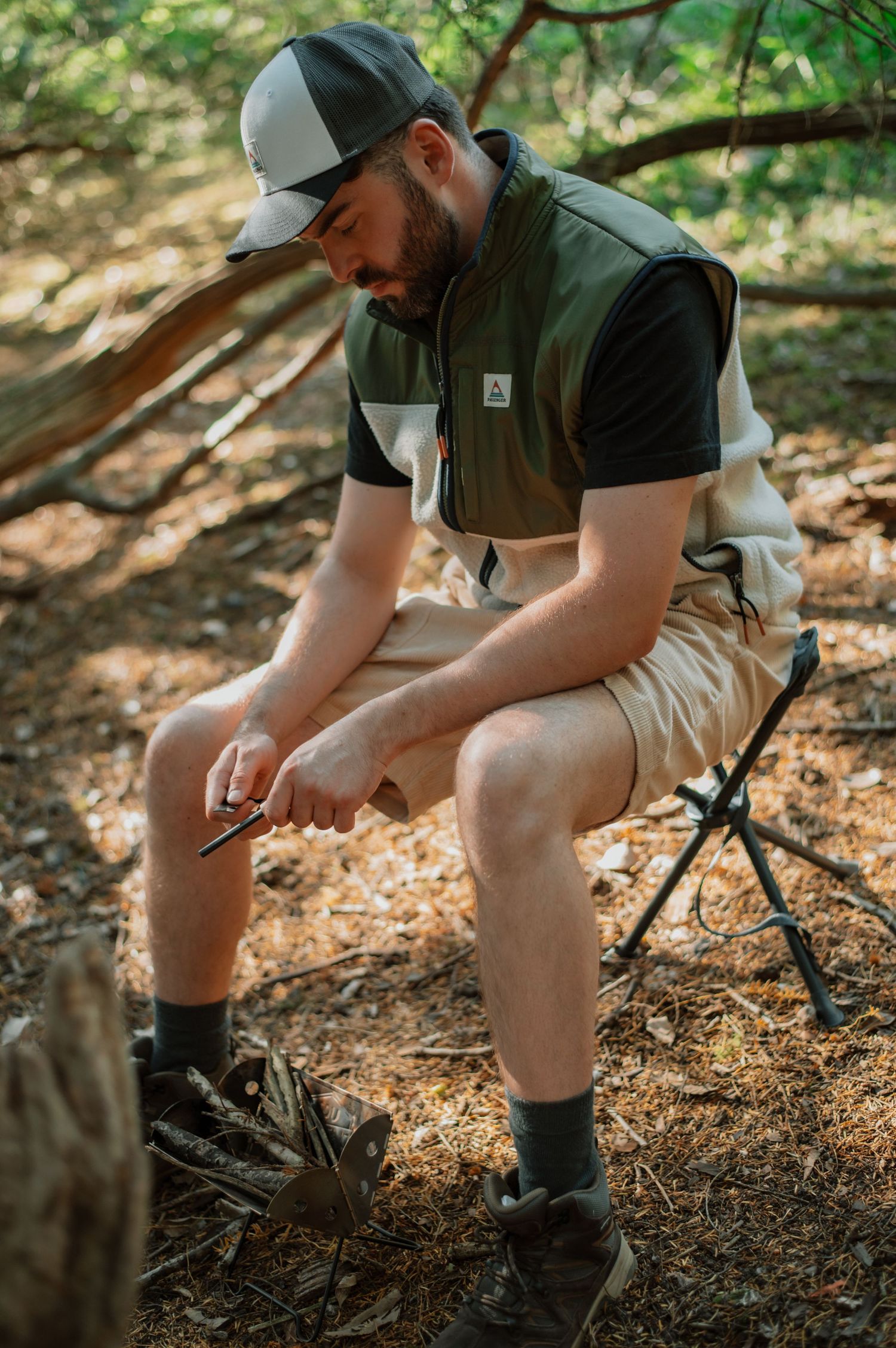 Man in olive vest and cap sits on camp stool in forest, examining small object in hands.