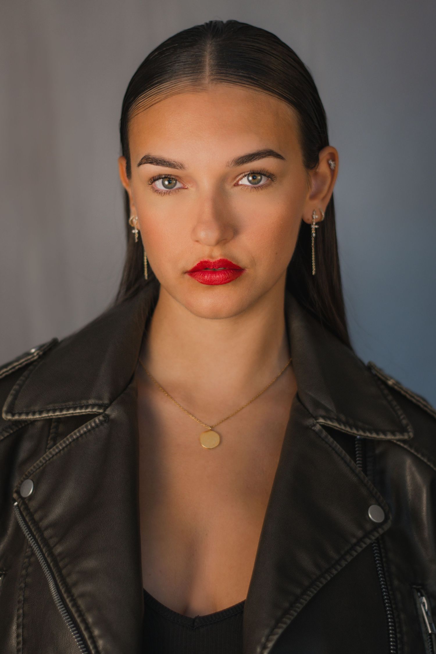 Edgy portrait in black leather jacket with bold red lips against moody gray background.