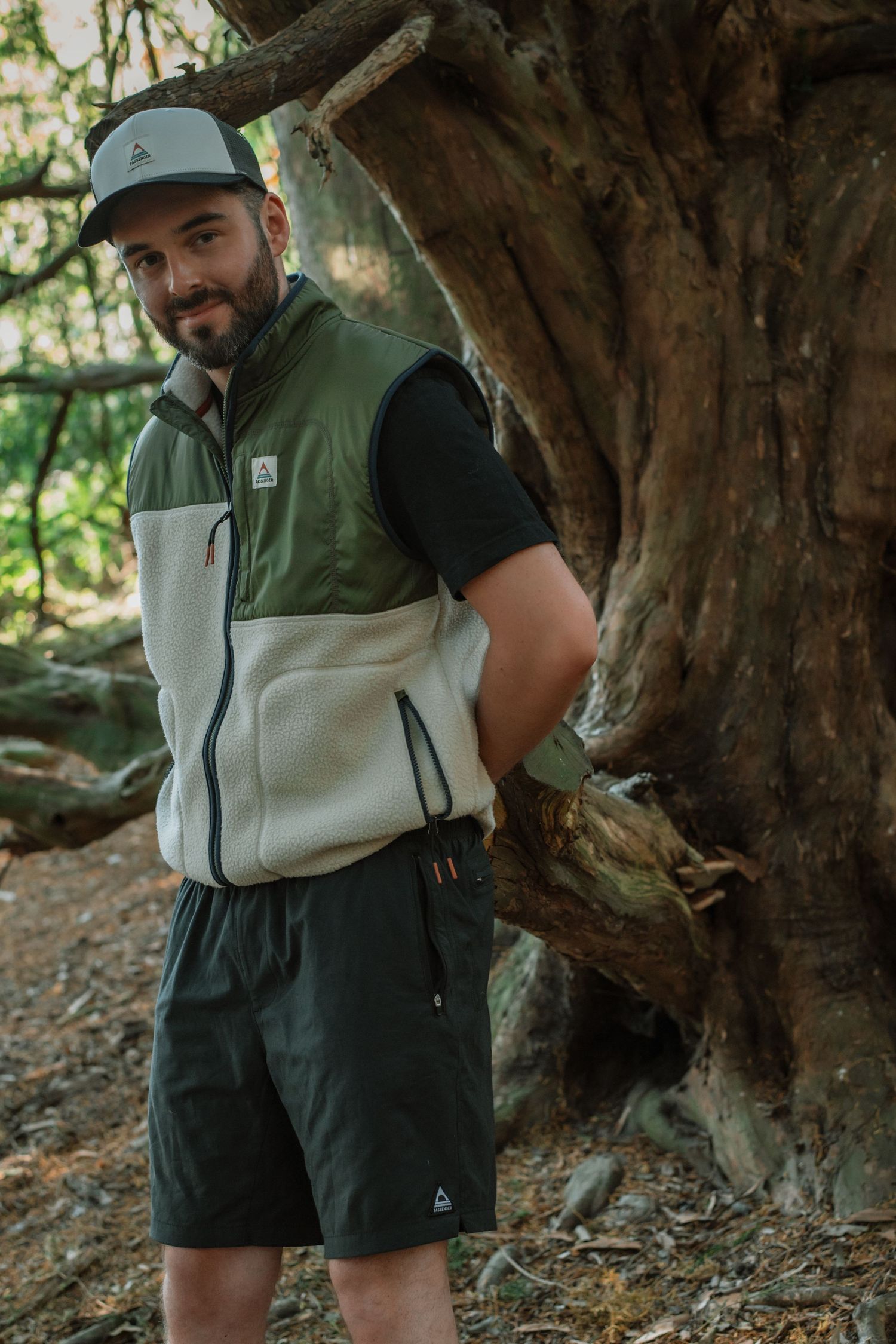 Man wearing green and cream fleece vest with shorts poses by a large tree in a forest setting.