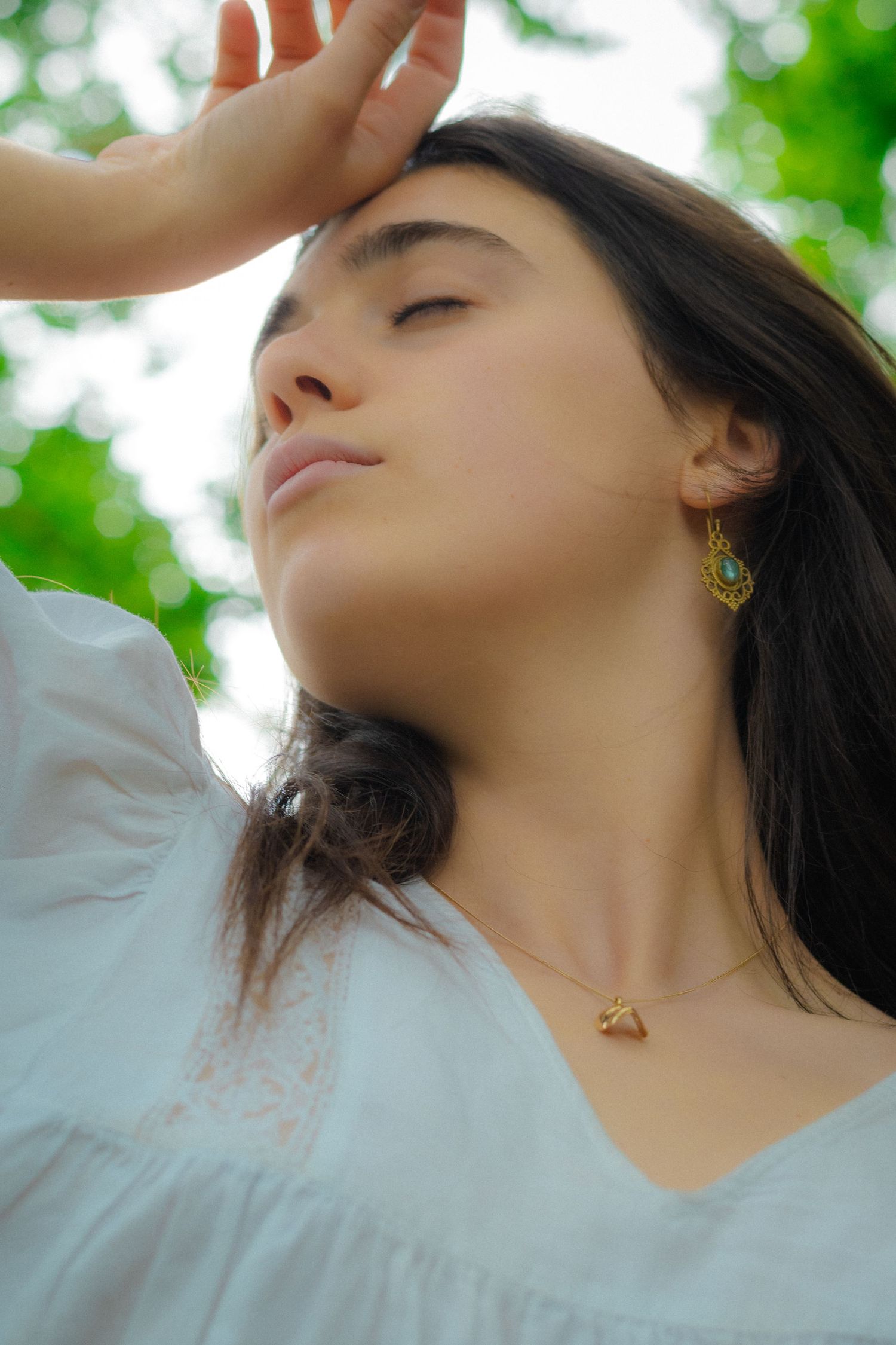 A woman with long dark hair and gold earrings gazes upward against a soft green foliage background in summer sunlight.