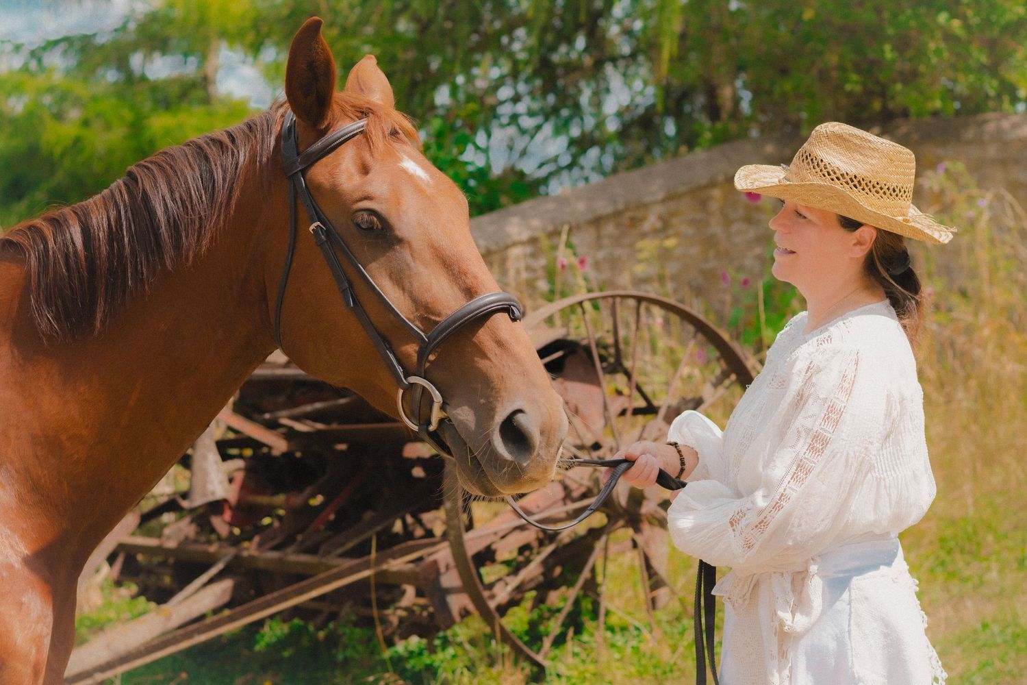 A brown horse stands beside someone wearing a white dress and straw hat in a sunny outdoor setting.